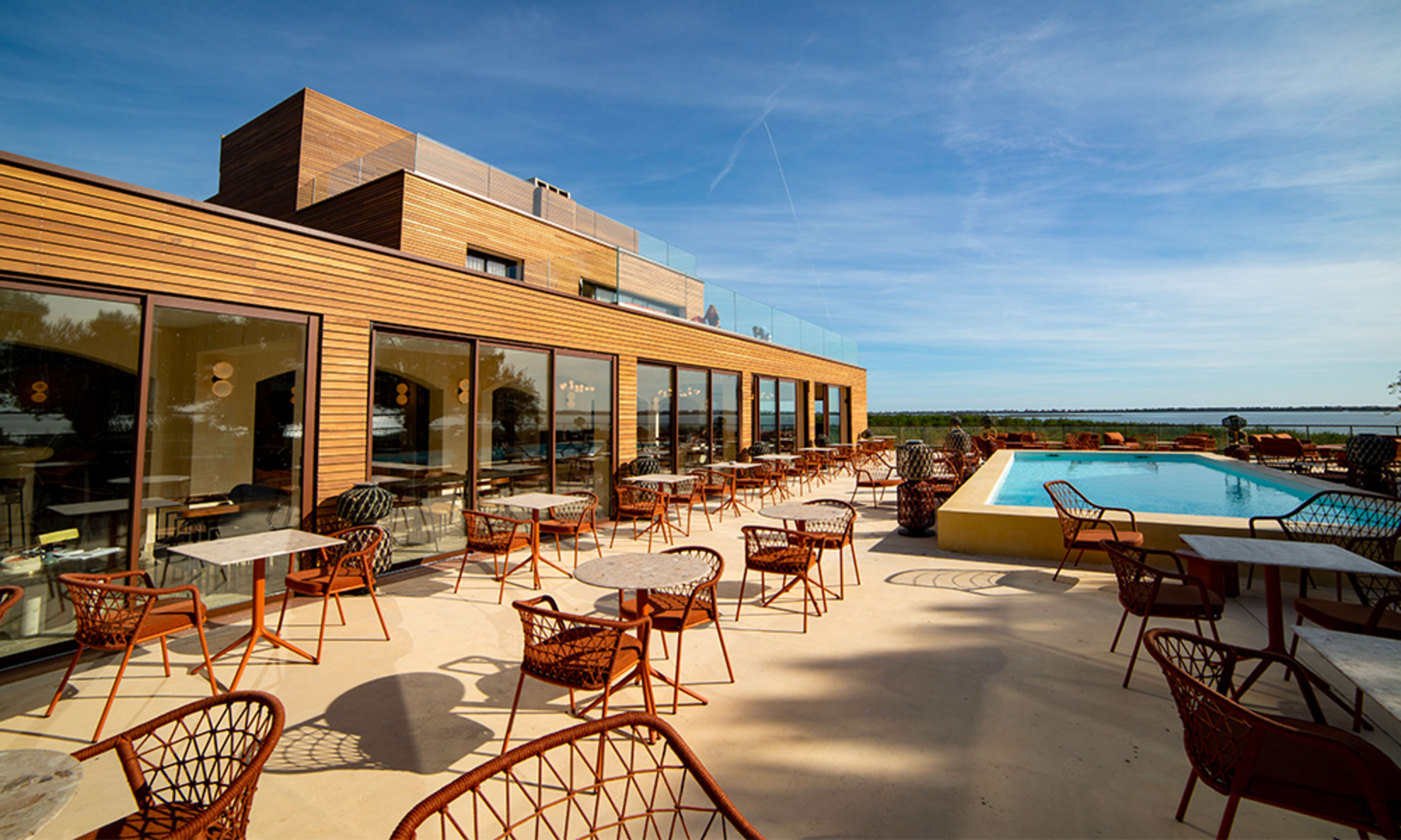 The image shows a modern building with large glass windows, a swimming pool, and outdoor seating arranged around tables on a sunlit patio overlooking water.