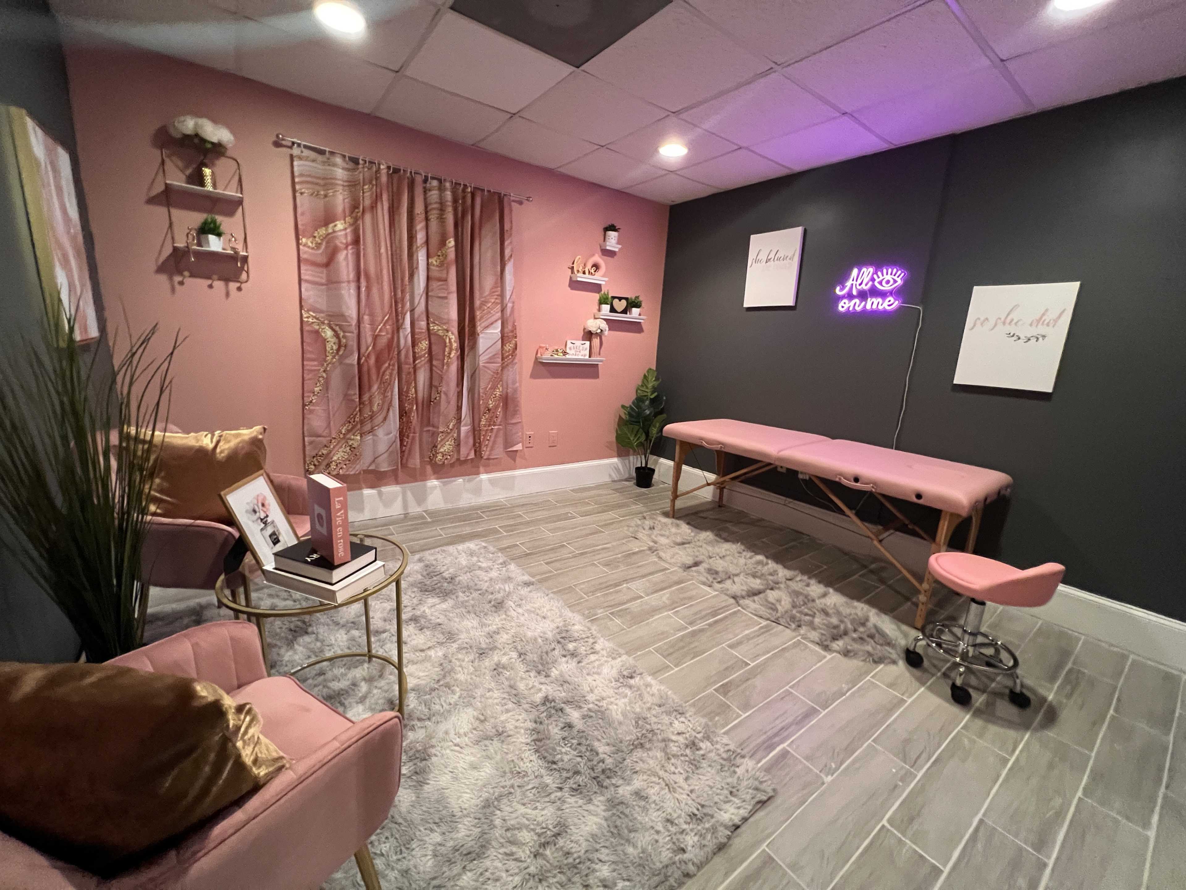 A neatly arranged beauty treatment room featuring a pink treatment table, two pink chairs, a rug, wall decor, and ambient lighting.