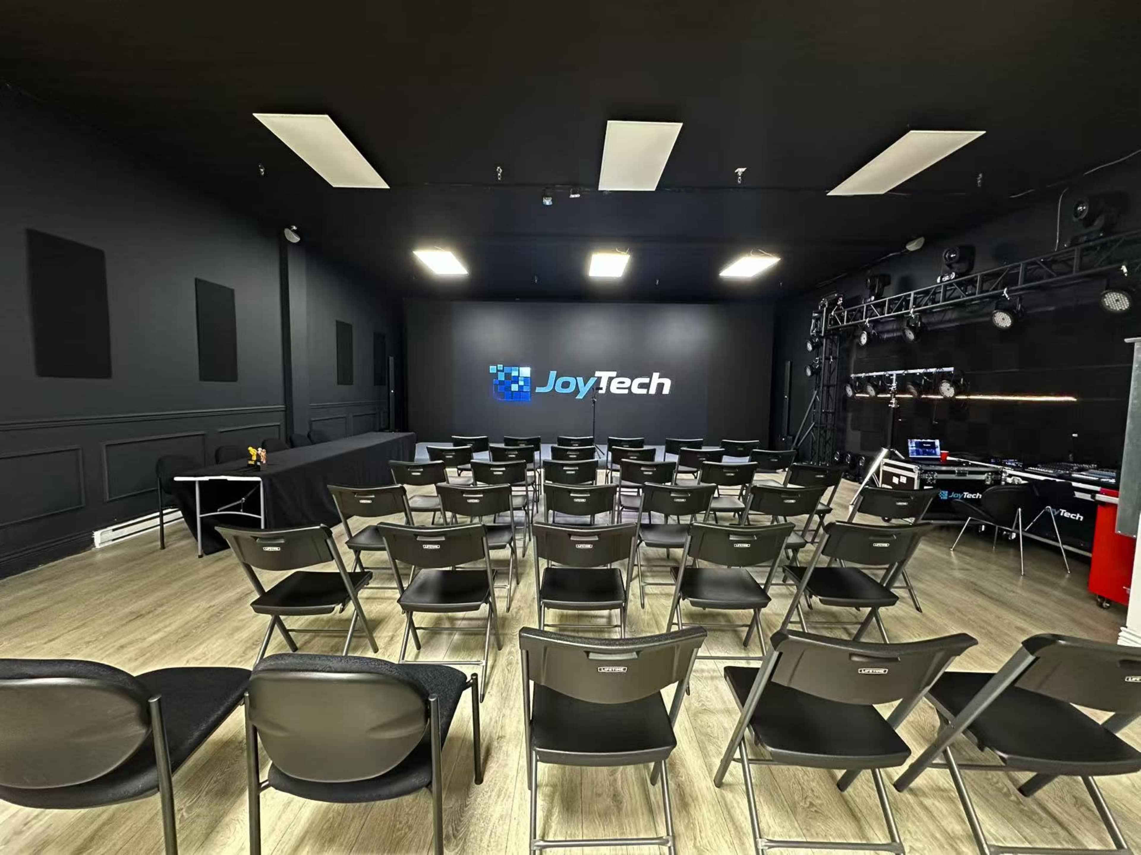 The image shows a conference room with rows of black chairs facing a large screen displaying the "JoyTech" logo, set against a dark wall.