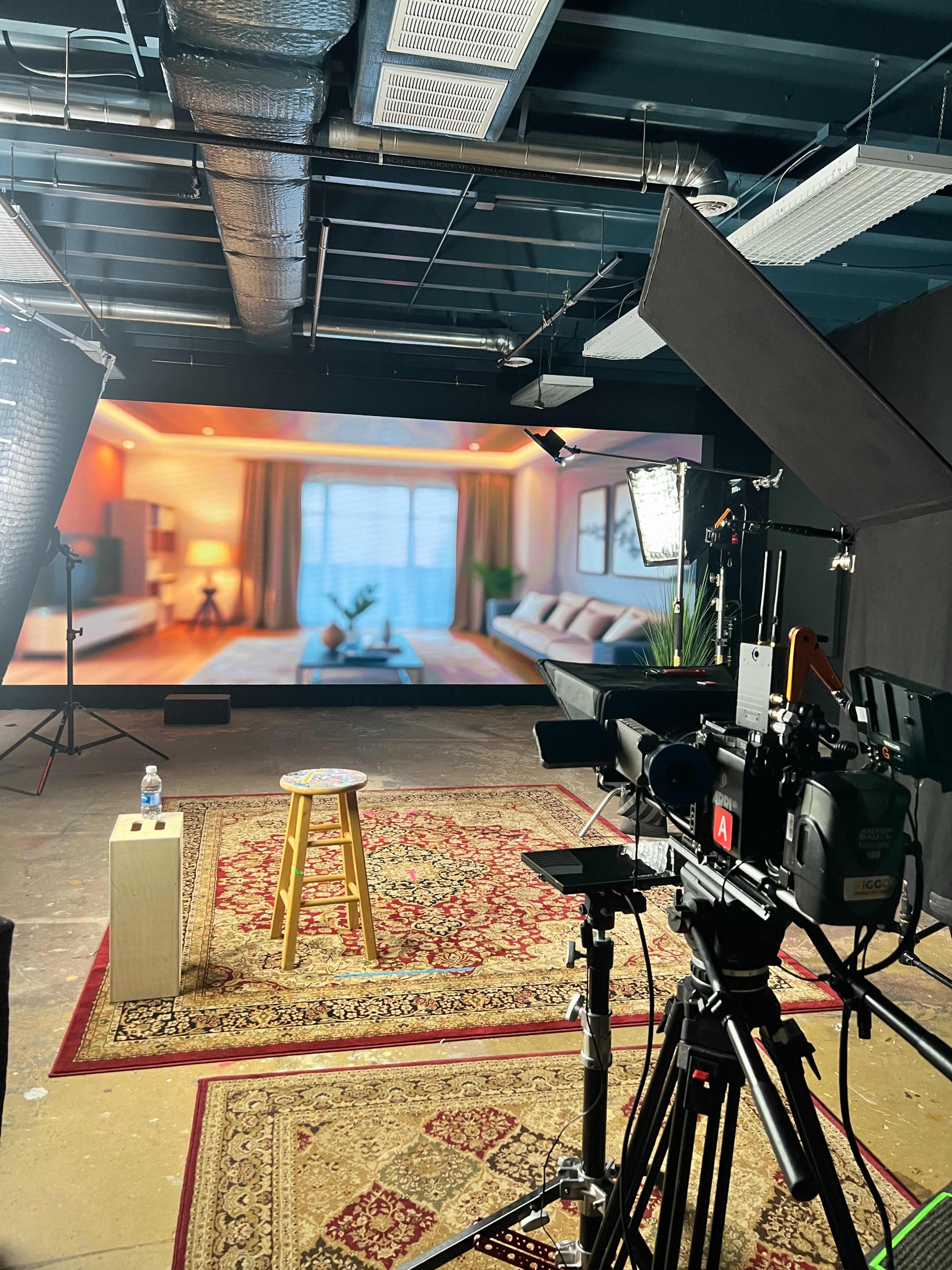 The image shows a film studio setup with a camera on a tripod, a stool, and a large screen displaying a living room scene in the background.