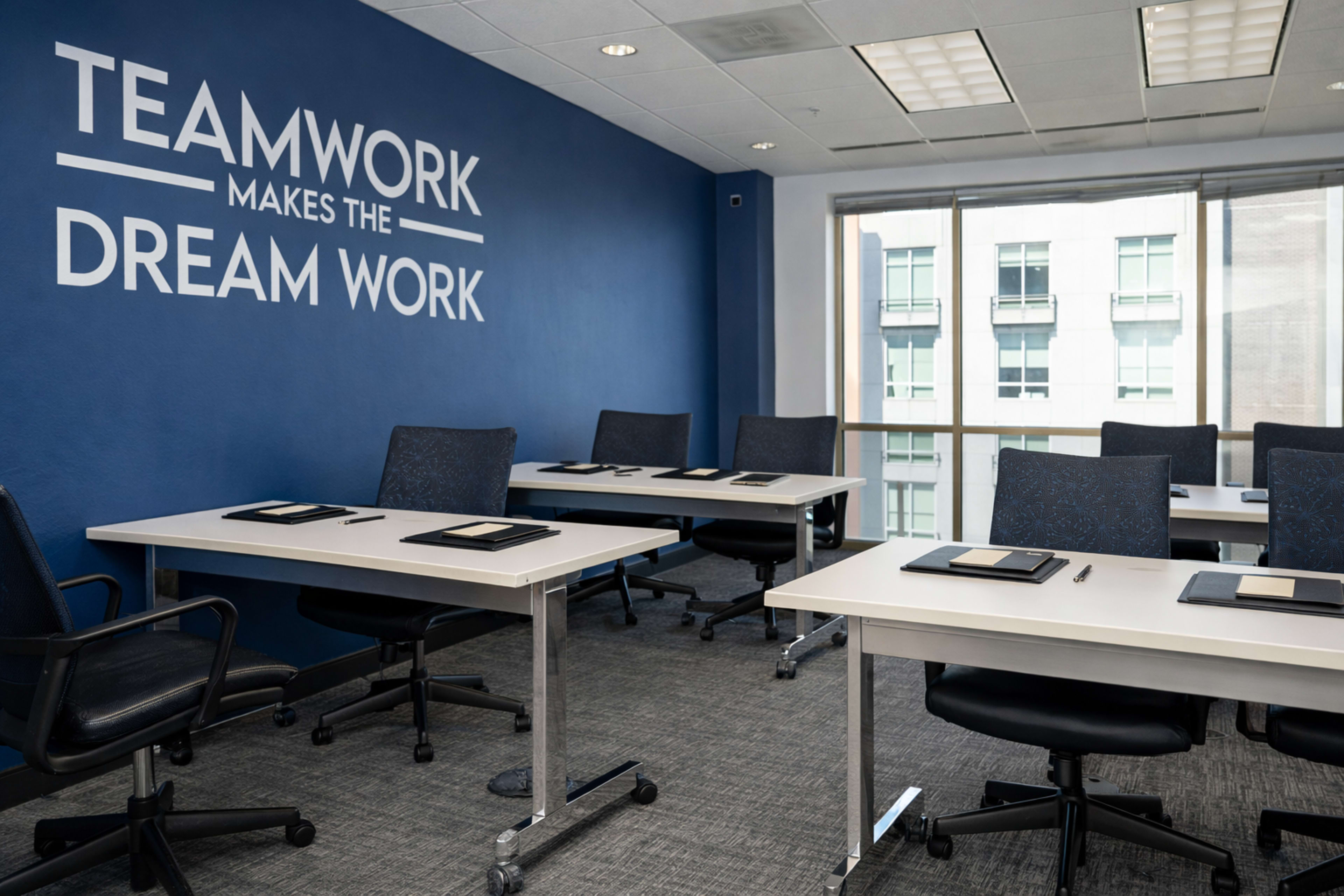A conference room features multiple tables and chairs arranged neatly against a blue wall with the phrase "TEAMWORK MAKES THE DREAM WORK" displayed.