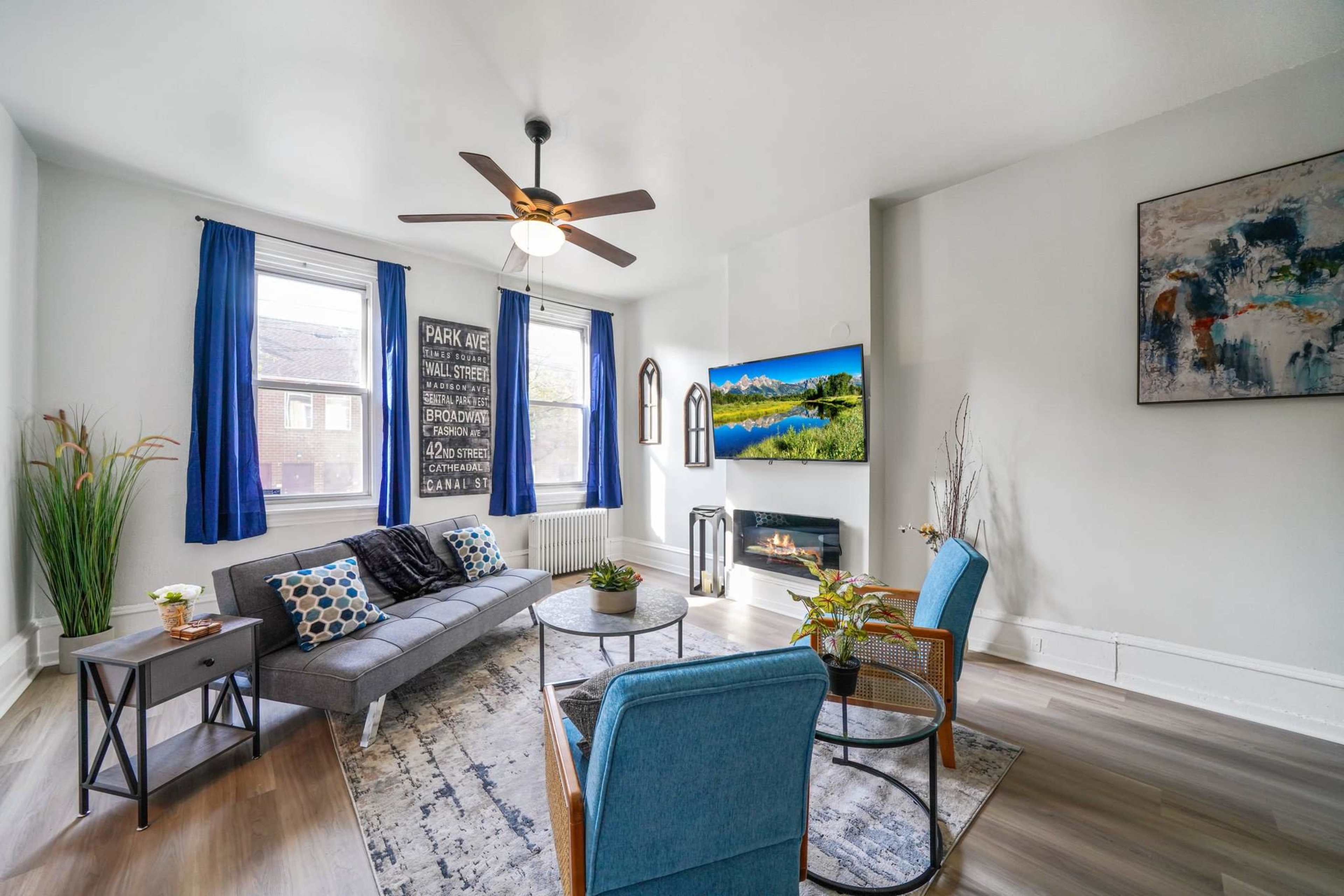 A modern living room featuring a gray sofa, blue accent chairs, and a large flat-screen TV mounted on a wall with a scenic nature view.