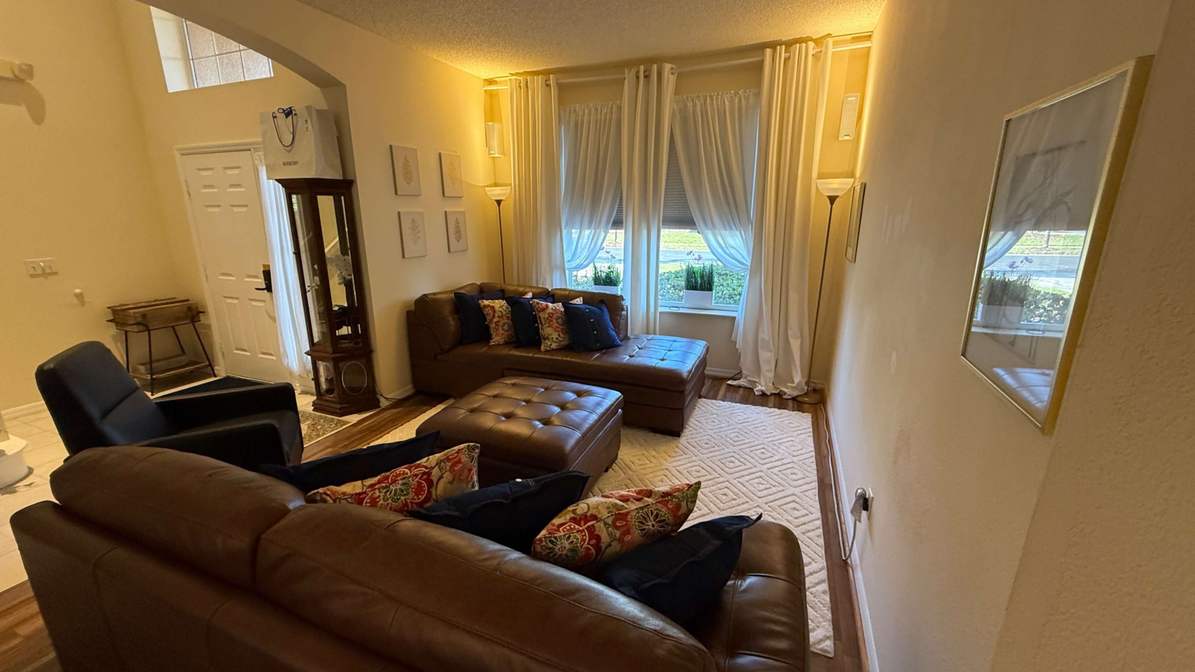 The image shows a living room with brown leather furniture, large windows adorned with sheer curtains, and decorative pillows on the sofa.