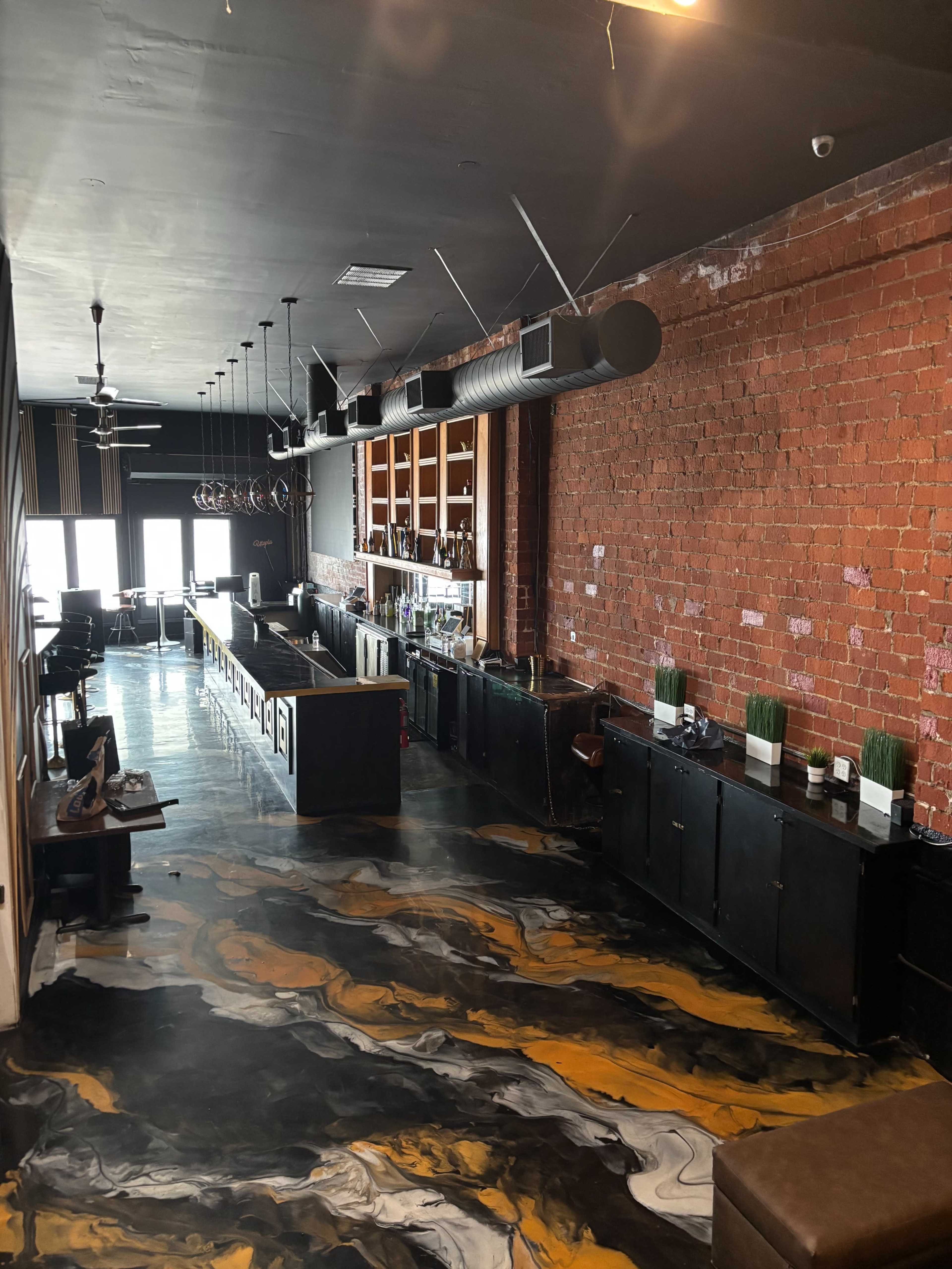 The image shows a modern bar area with a long counter, exposed brick walls, and a decorative marbled floor.