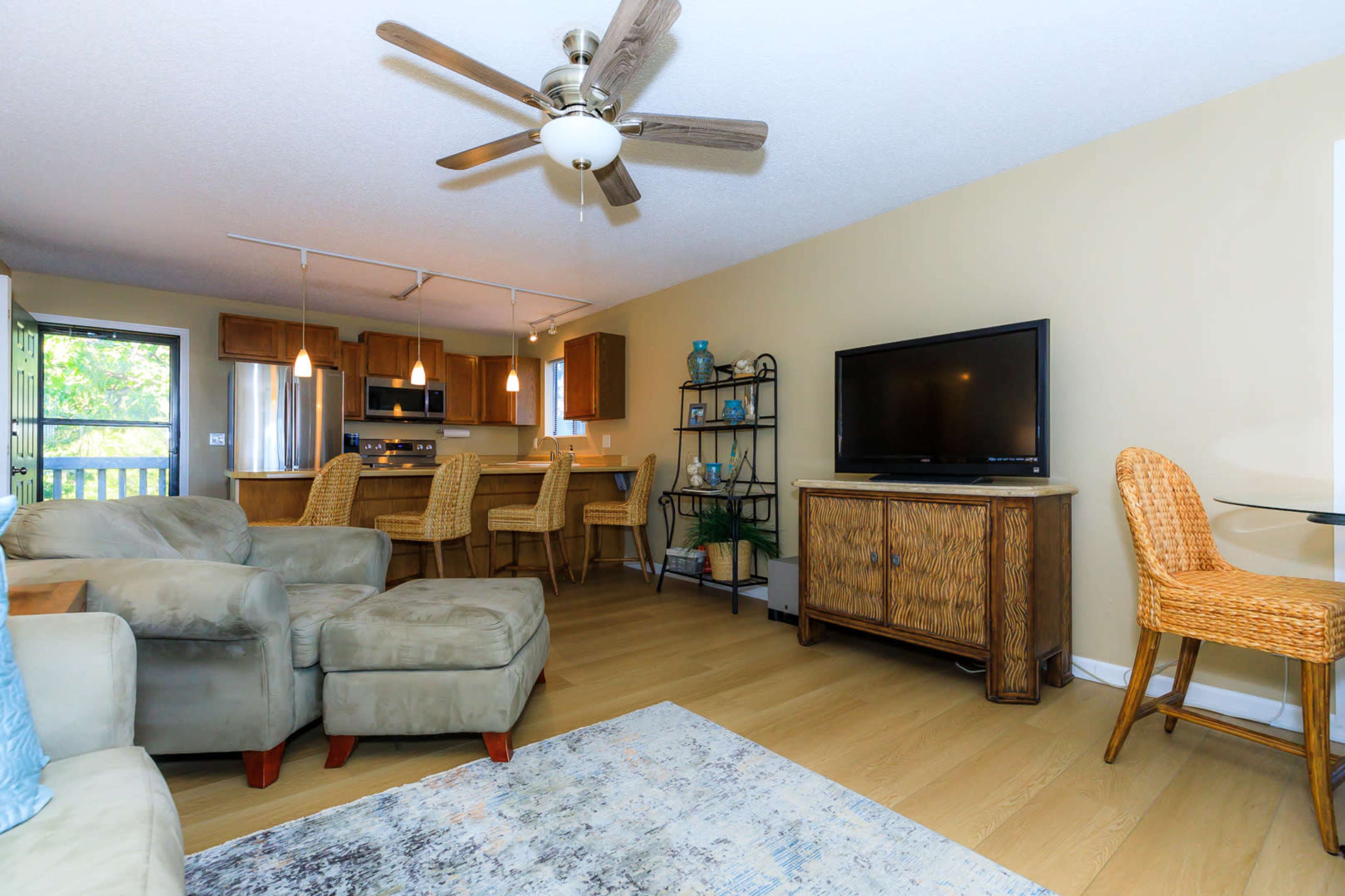 The living area features a ceiling fan, a television on a cabinet, and beige furniture, opening up to a kitchen and dining area in the background.