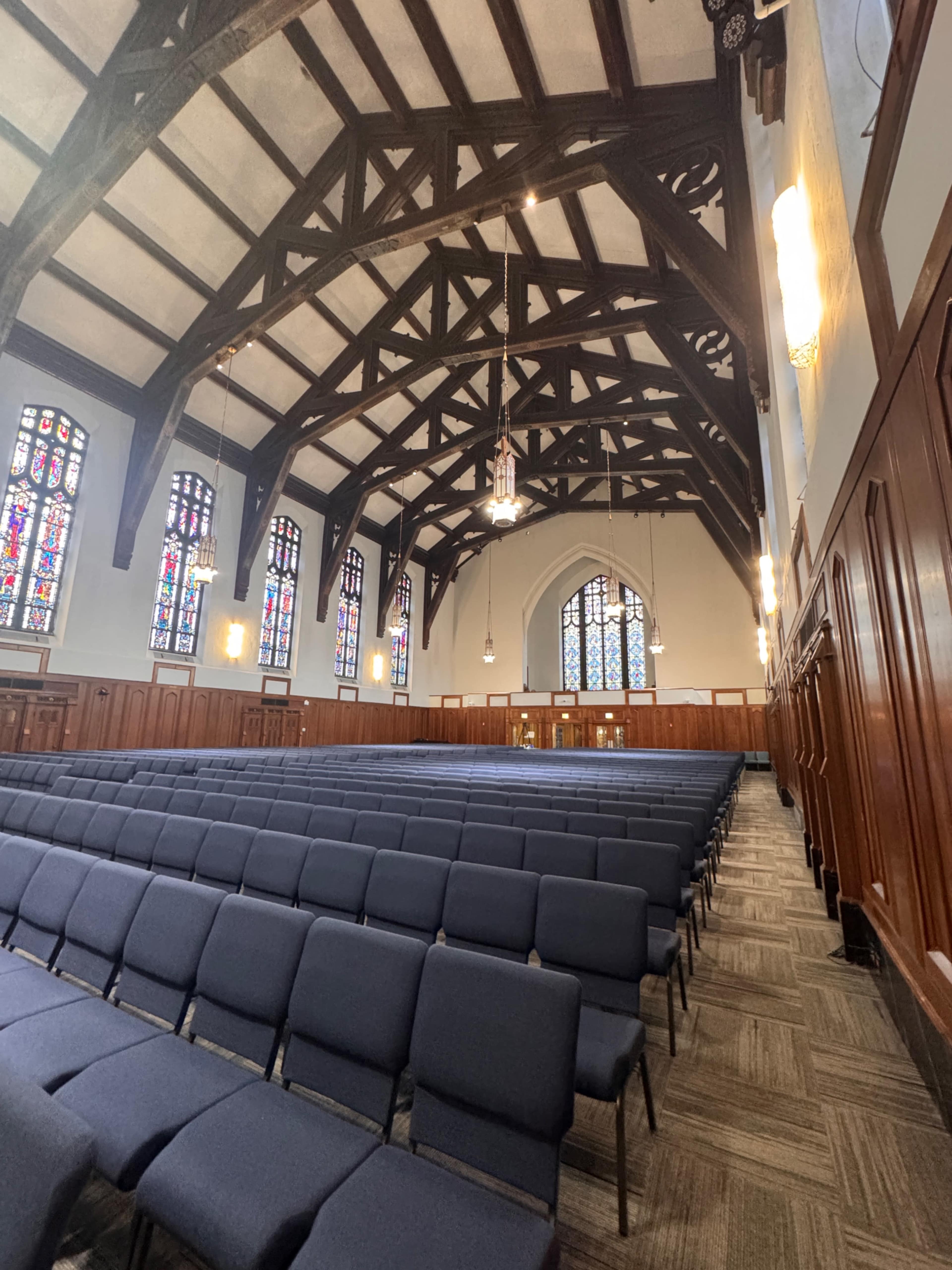 The image shows an interior view of a church with wooden beams, stained glass windows, and rows of blue chairs arranged for an event.