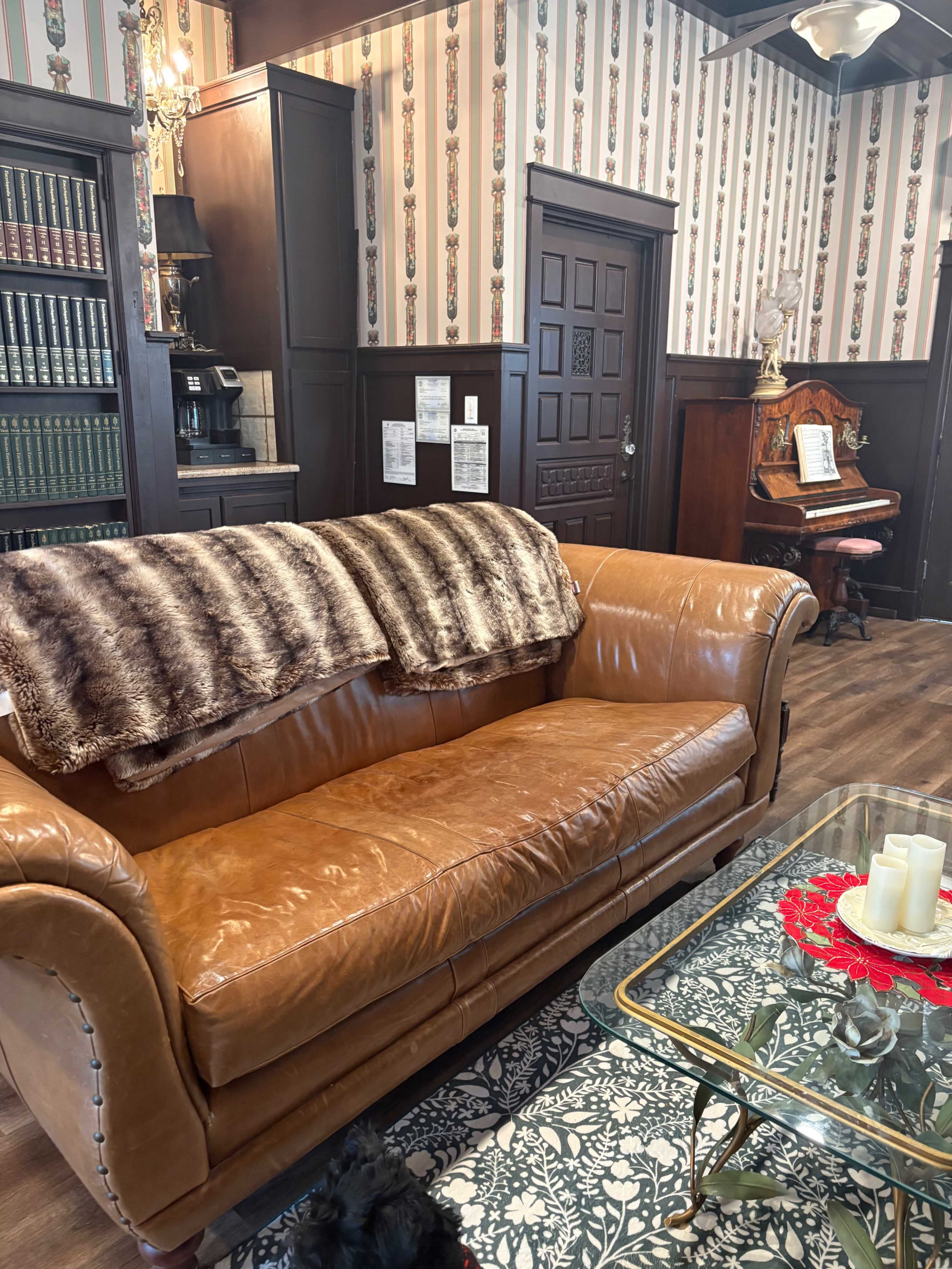 The image shows a cozy room with a brown leather couch, a glass coffee table, and vintage furnishings, including a wooden piano and a patterned wallpaper.