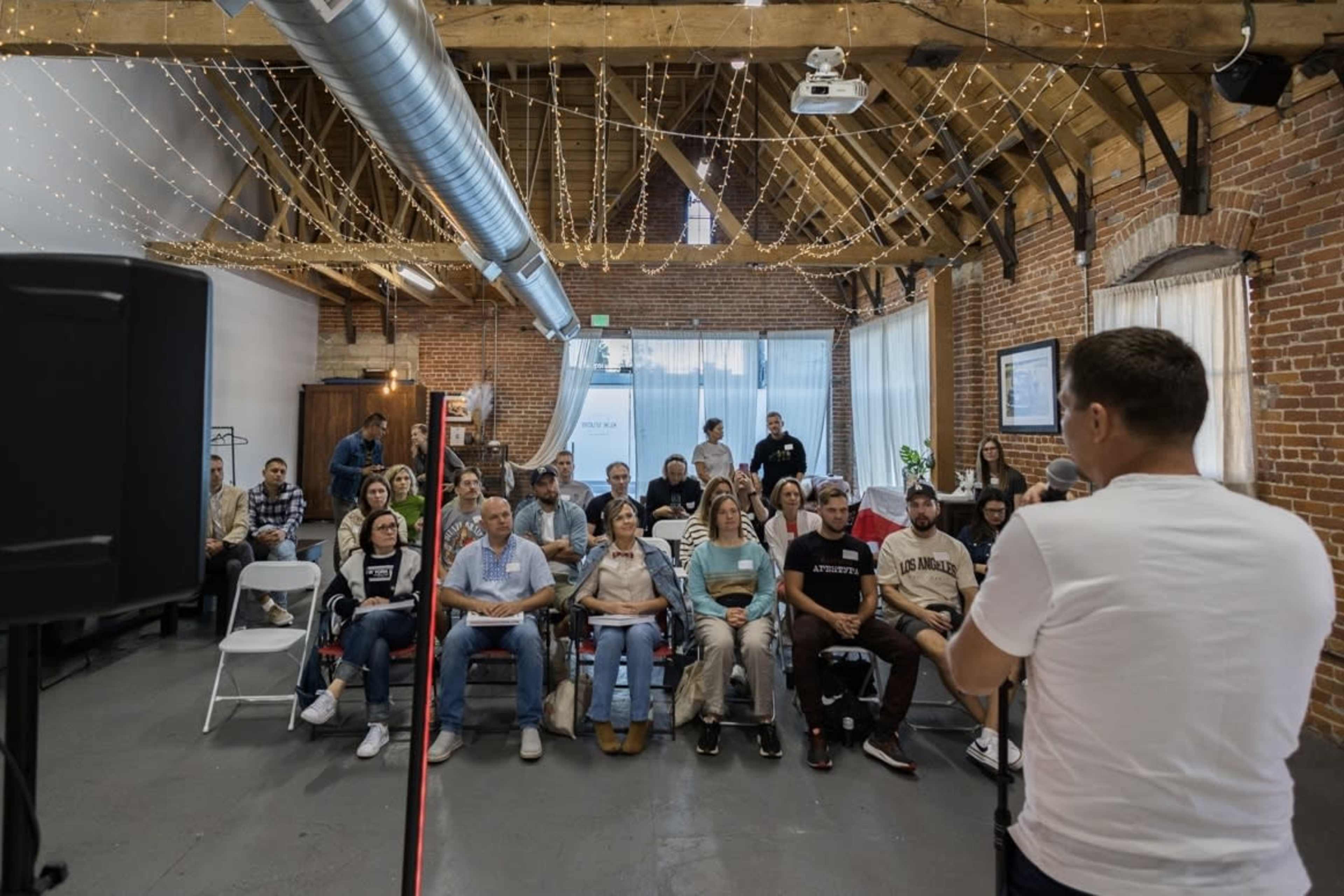 A speaker addresses a seated audience in a room with exposed brick walls and string lights overhead.