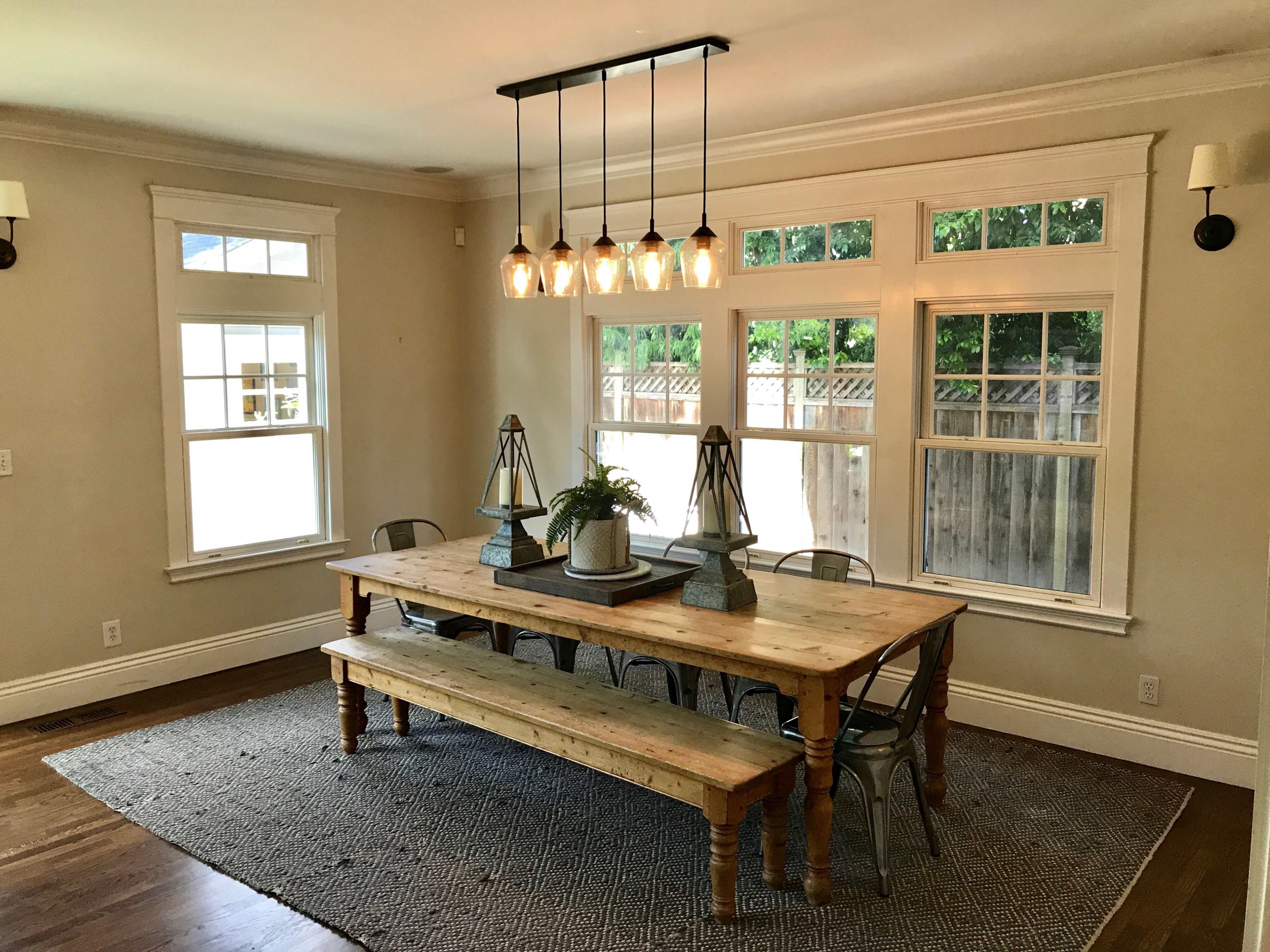 A large wooden dining table is centered in a well-lit room with multiple windows and a pendant light overhead, surrounded by metal chairs and decorative elements.