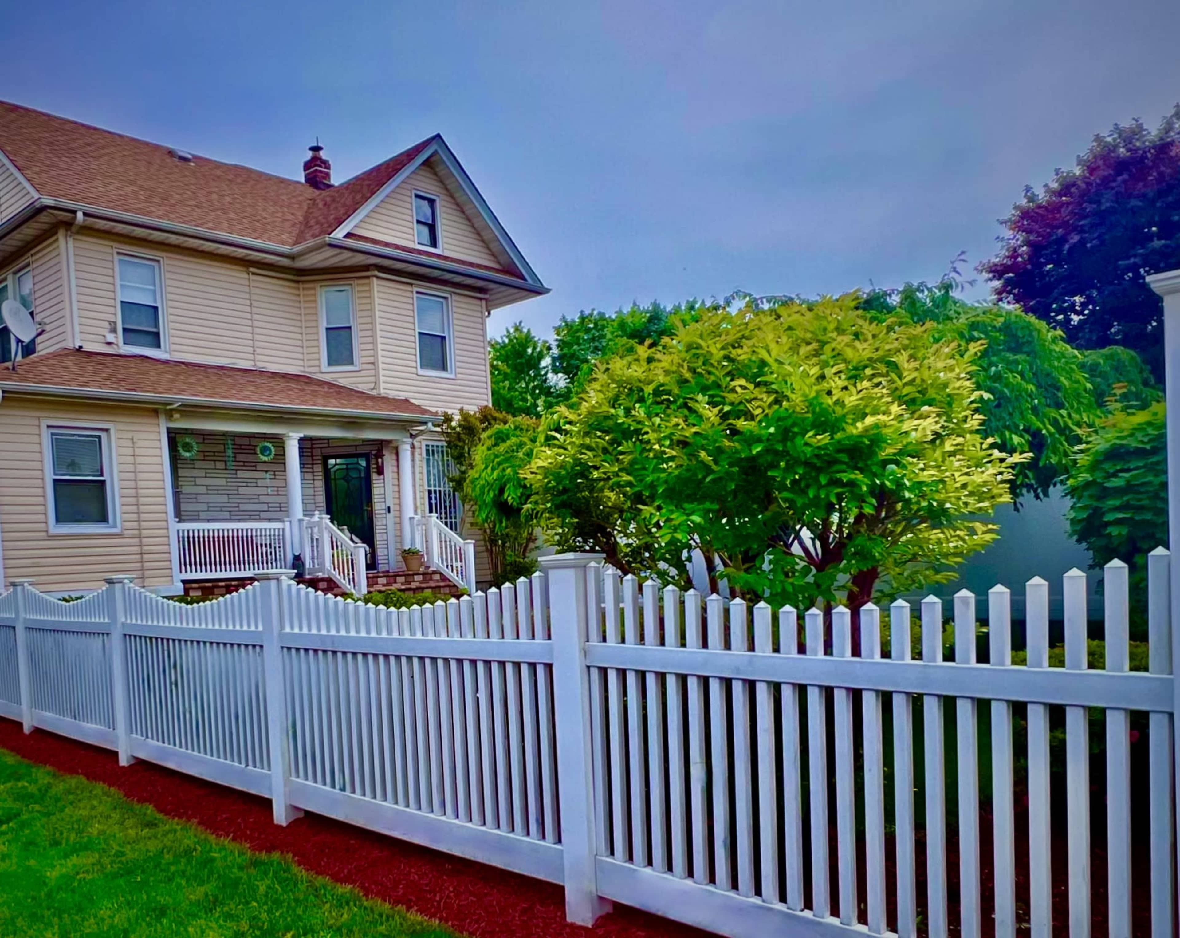 A two-story house with a light-colored exterior and a brown roof is enclosed by a white picket fence and surrounded by greenery.