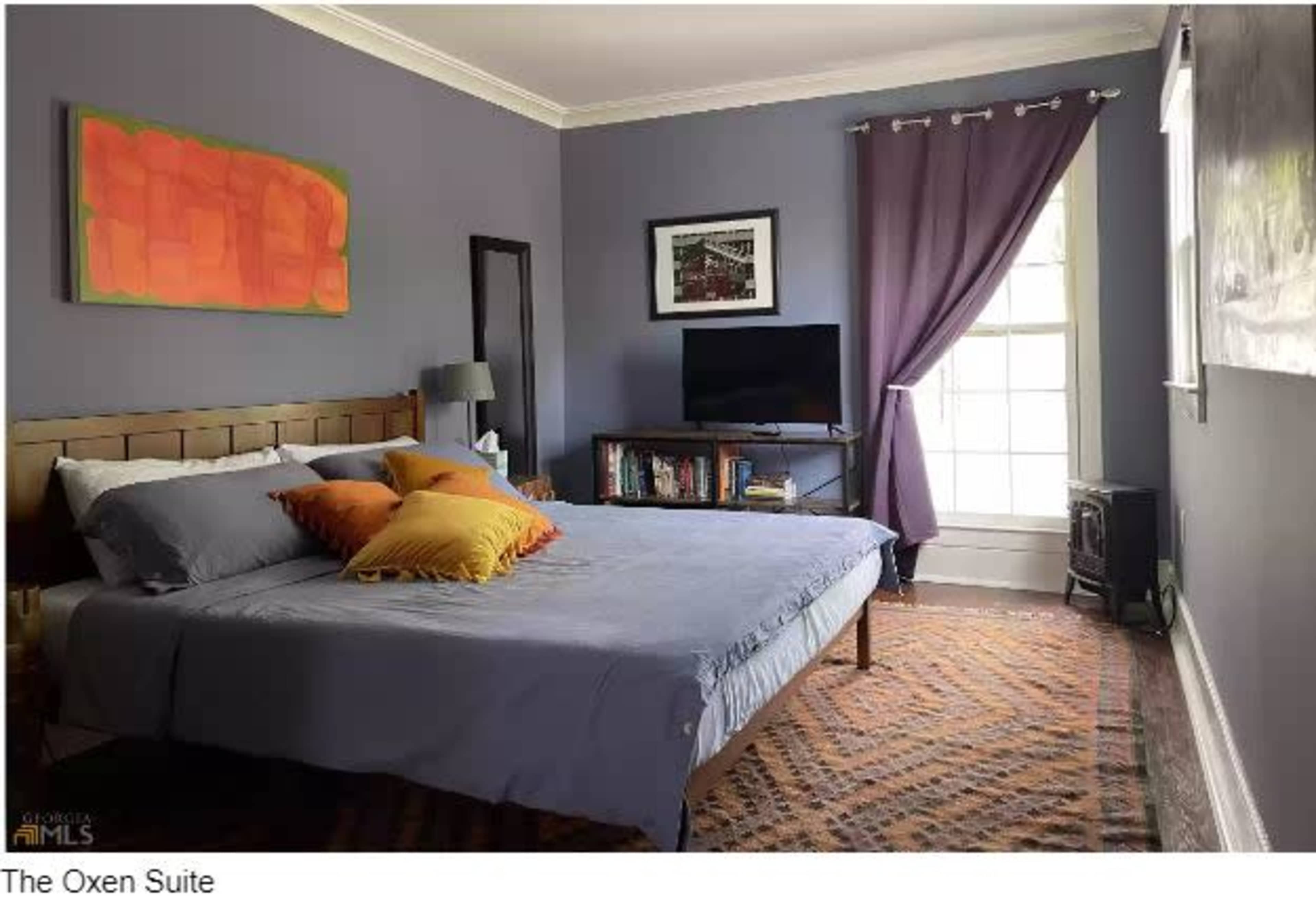 The image shows a bedroom with a bed, colorful pillows, a TV on a stand, curtains, and a window providing natural light.