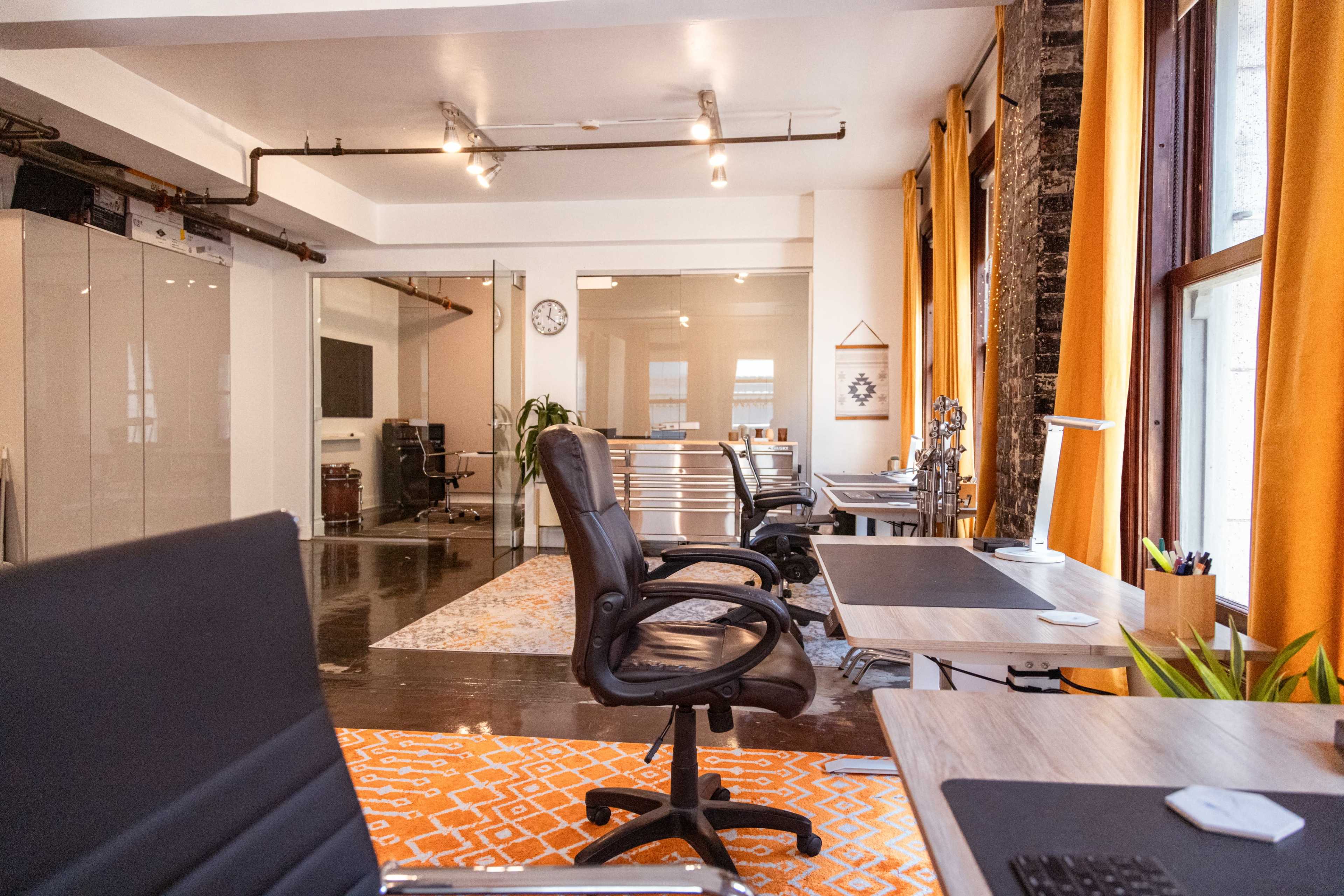 The image shows a modern office space with multiple workstations, ergonomic chairs, and large windows with orange curtains.