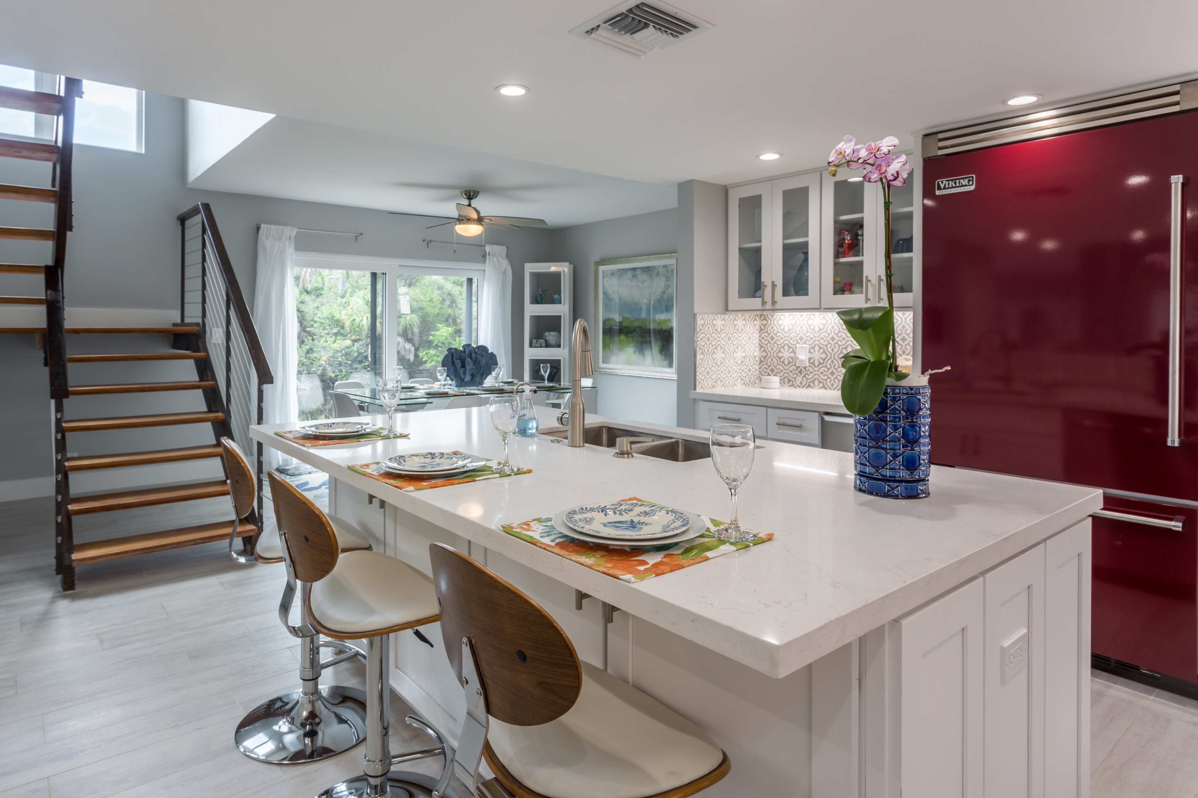 A modern kitchen features white cabinetry, a large island with seating, and a red refrigerator, accompanied by a staircase in the background.