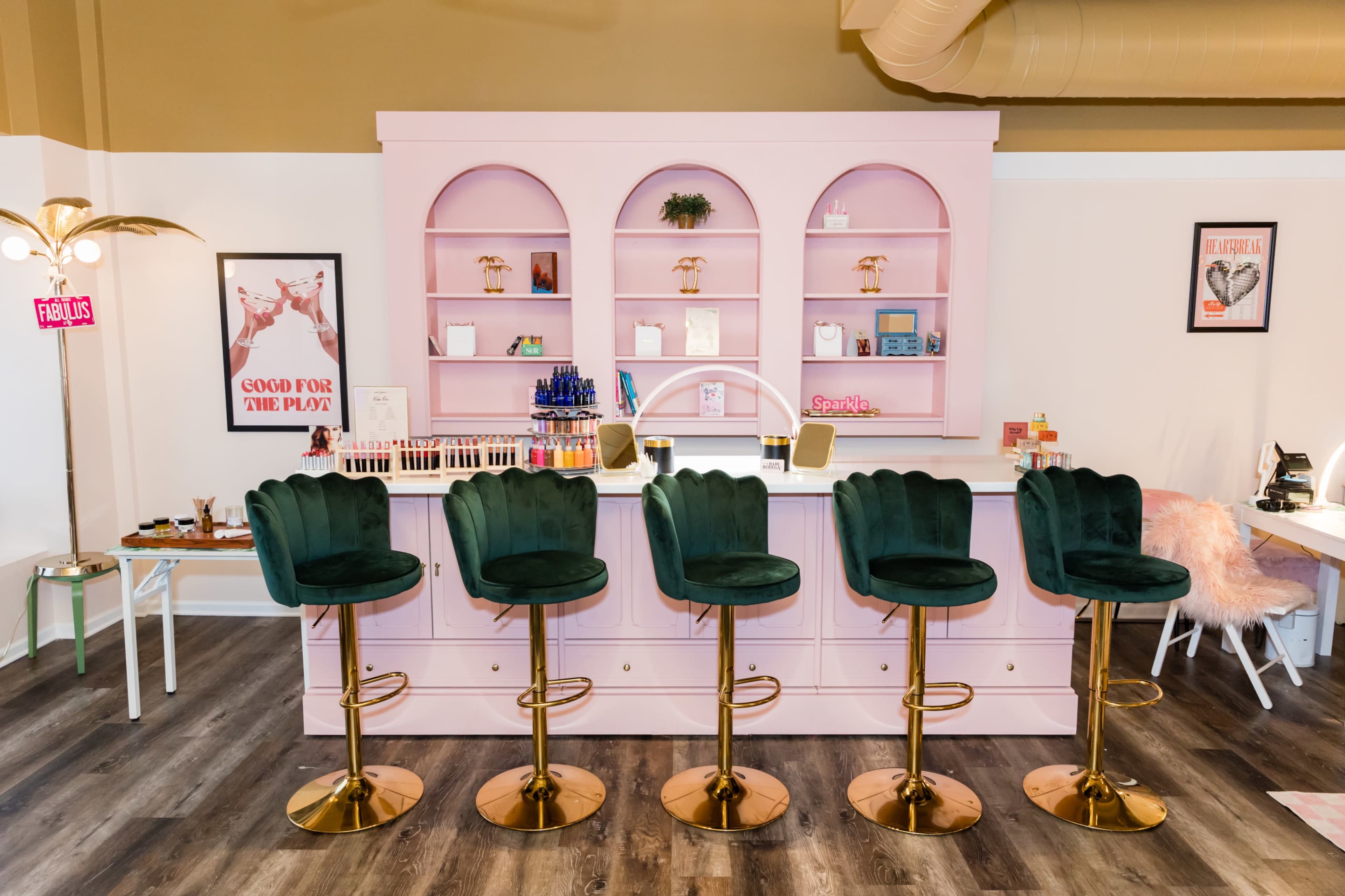A brightly lit beauty bar with a pink countertop and five green velvet barstools.