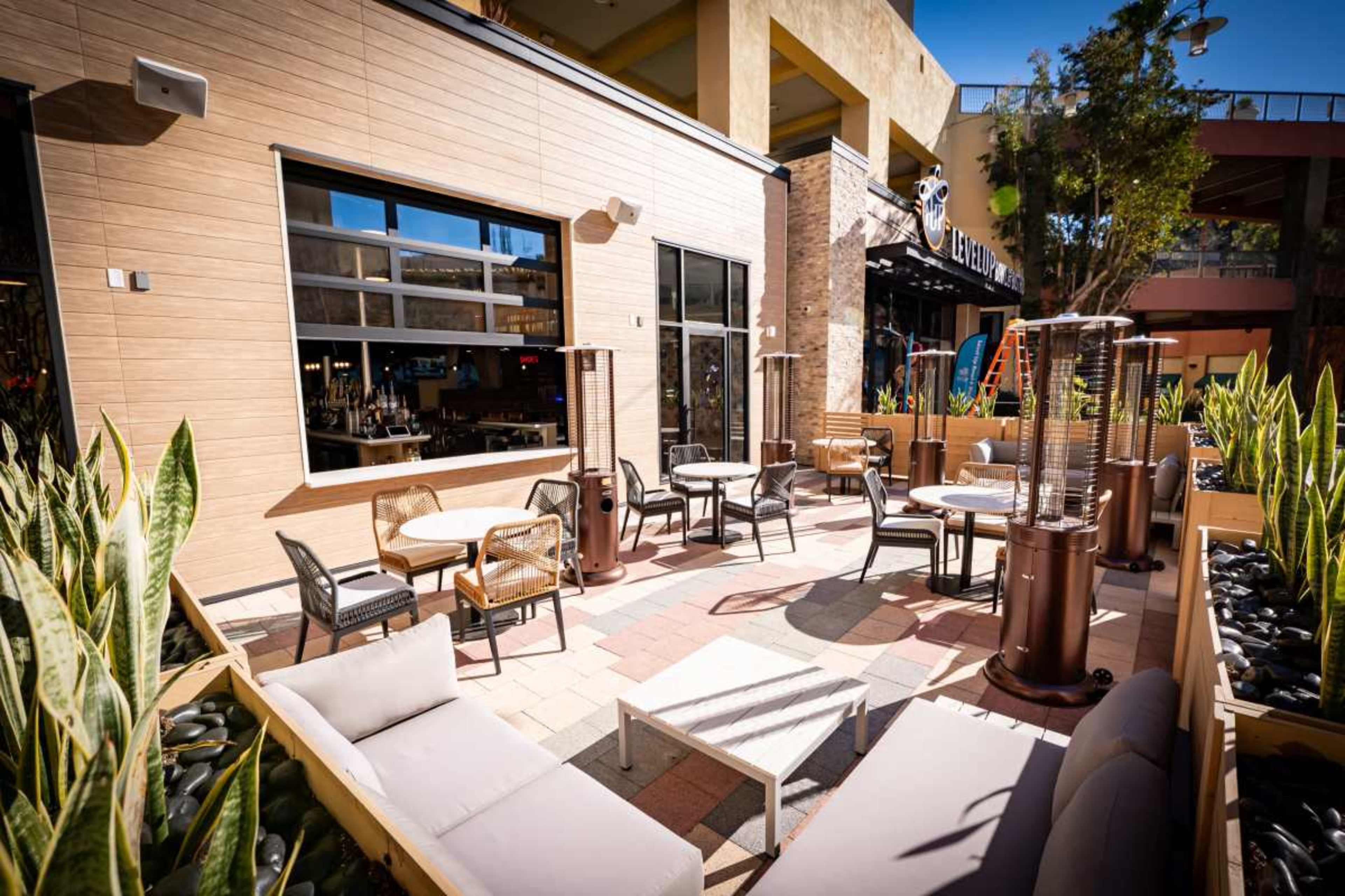 The image shows an outdoor seating area of a restaurant with tables, chairs, and patio heaters surrounded by decorative plants.