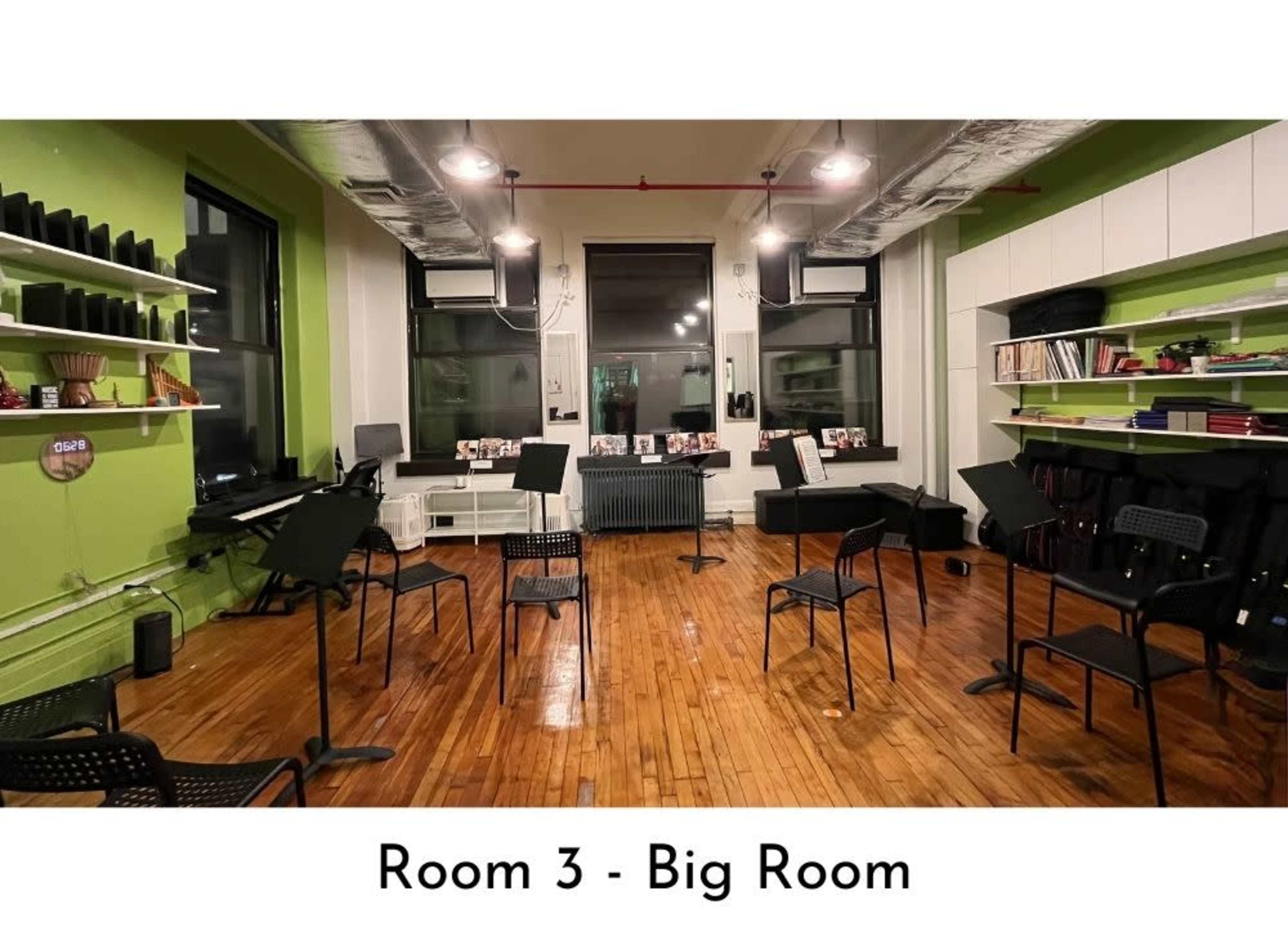 The image shows a spacious room with wooden floors, several black chairs arranged around music stands, and green walls with shelves holding books and other items.