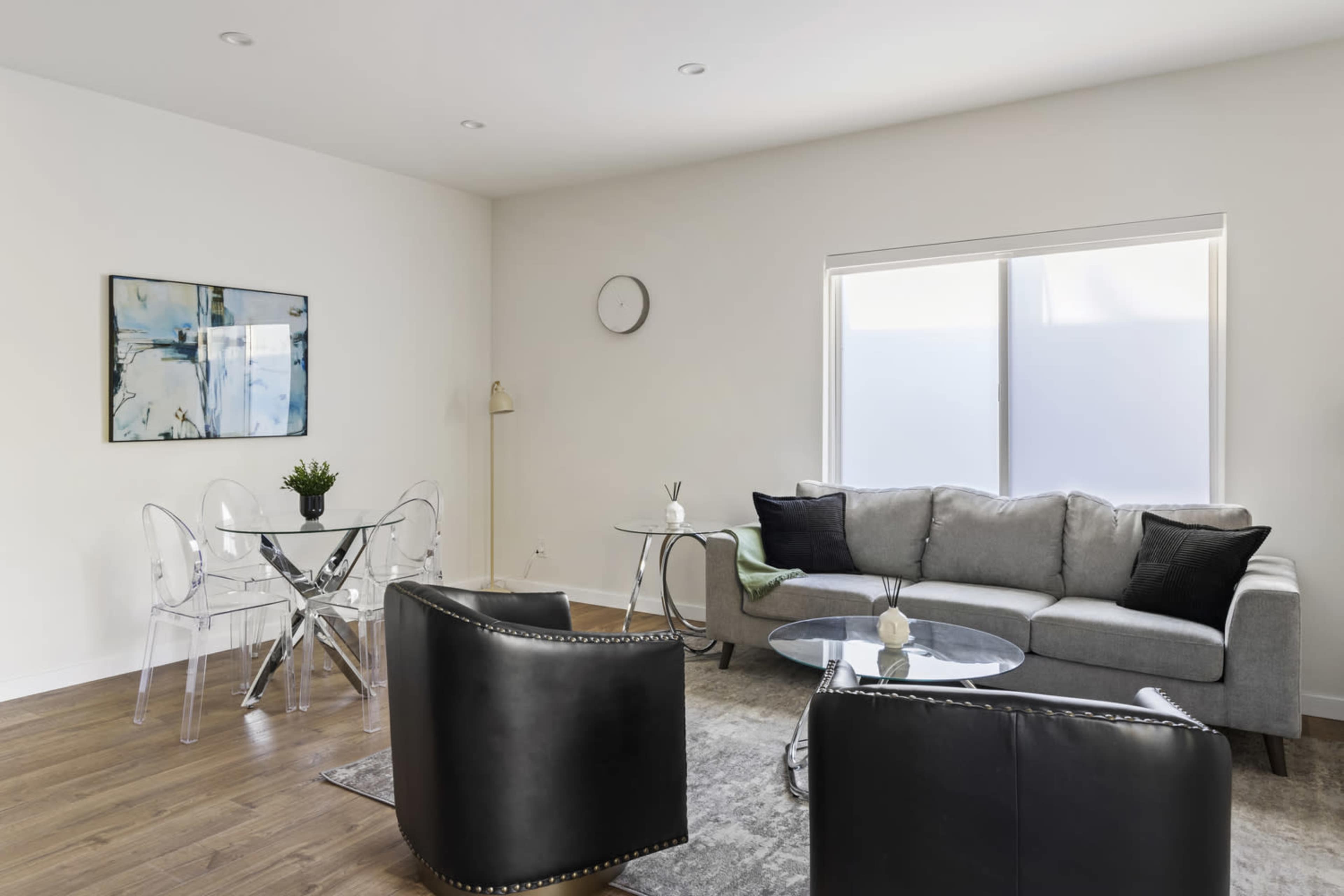 The image shows a modern living room featuring a gray sofa, two black armchairs, a glass coffee table, and a dining area with a round glass table.
