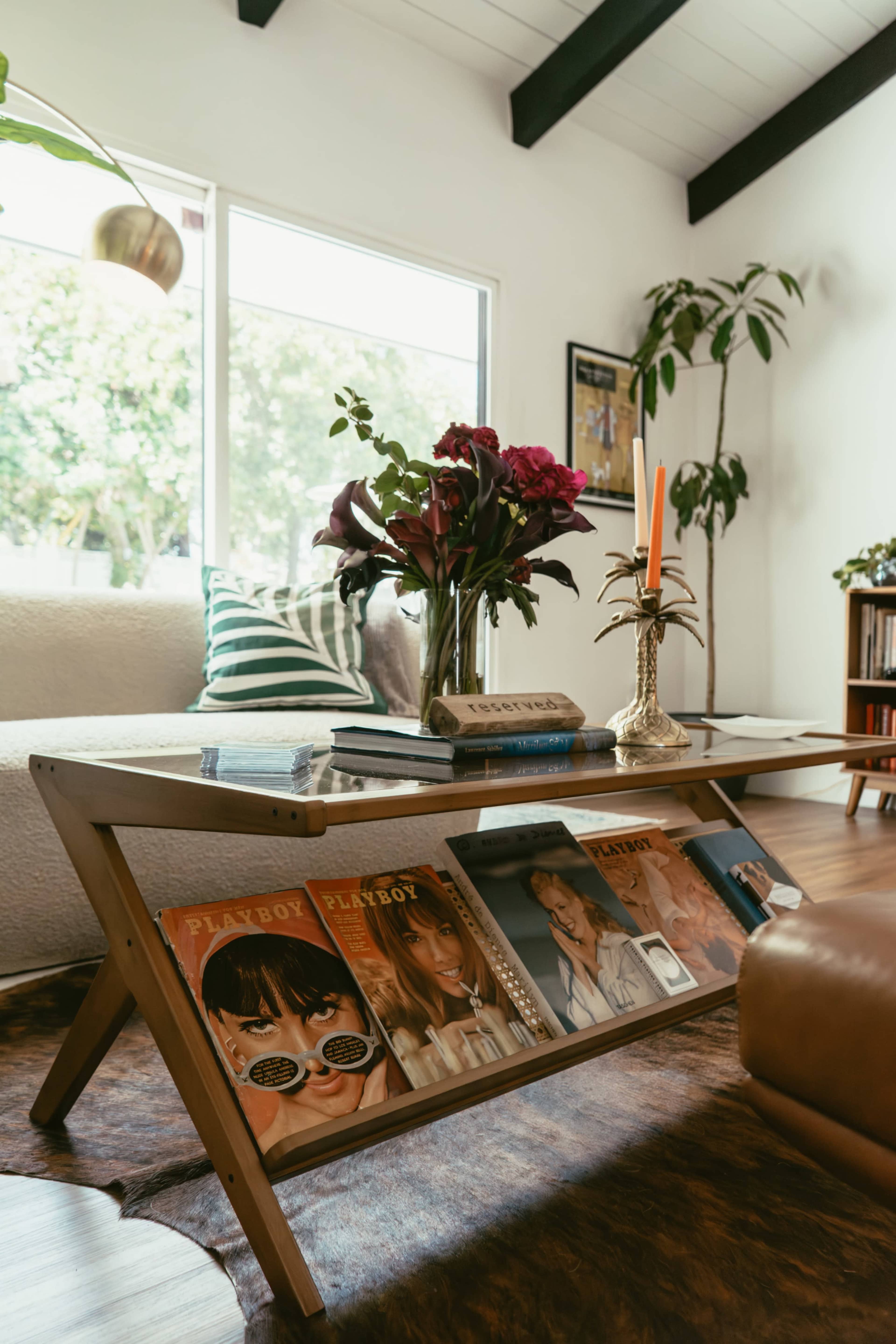 A modern living room features a wooden coffee table displaying vintage magazines, a vase of flowers, and a candle, with a sofa and plants visible in the background.
