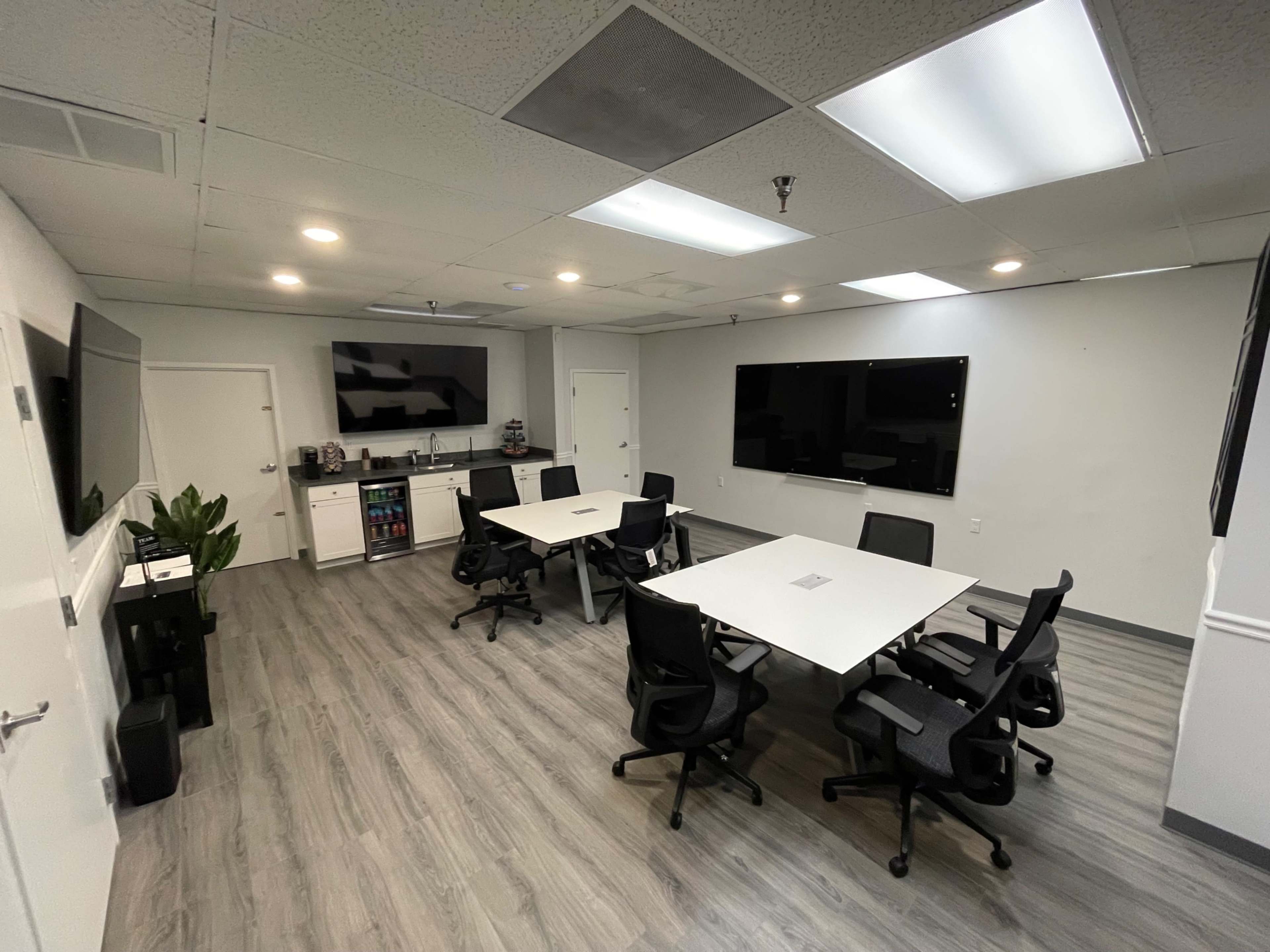 A modern conference room featuring multiple tables and chairs, two large screens on the walls, and a kitchenette area.