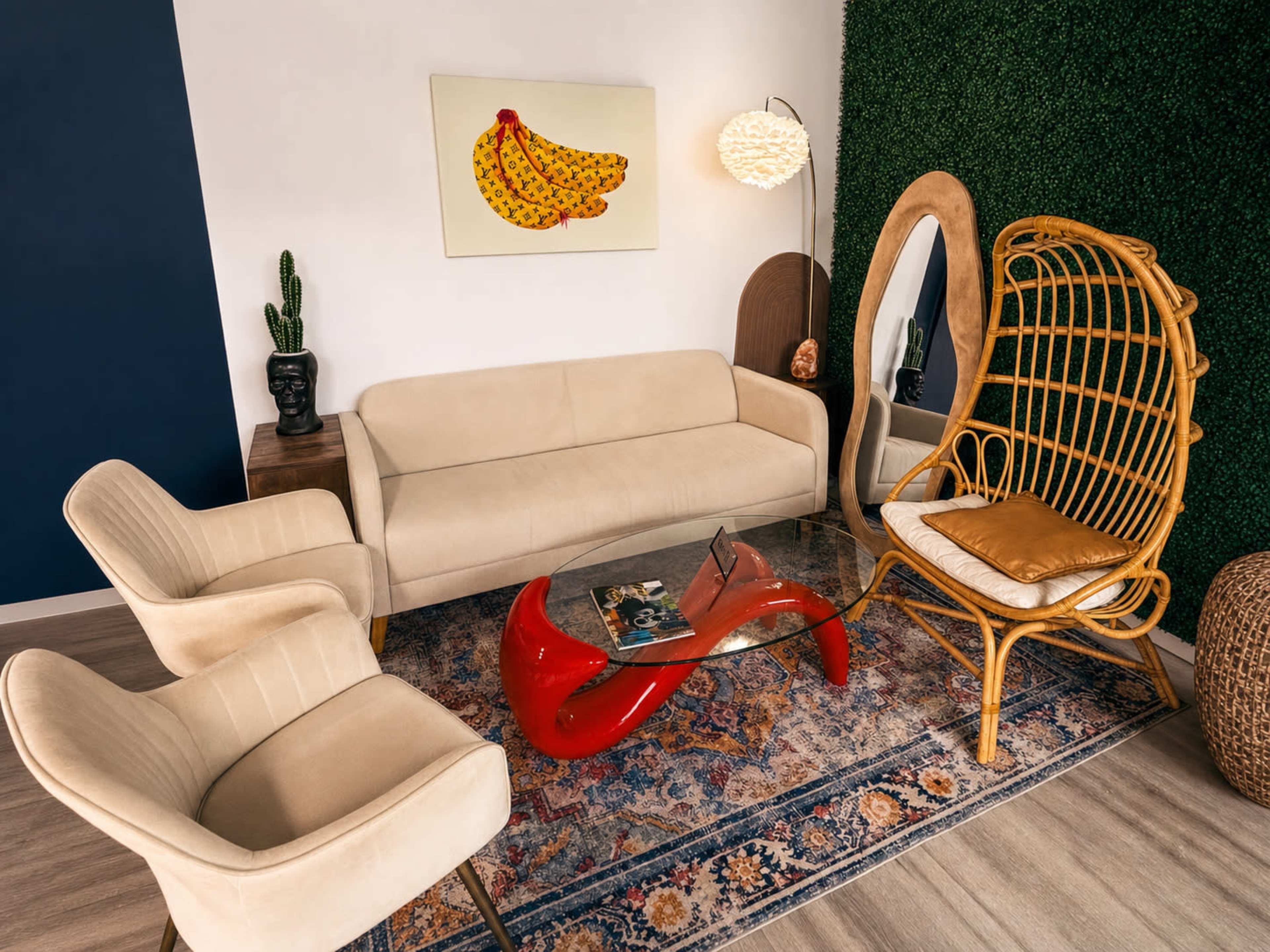 The image shows a modern living space featuring a beige sofa, two cream armchairs, a unique red coffee table, a rattan chair, and decorative elements like a banana painting and plants.
