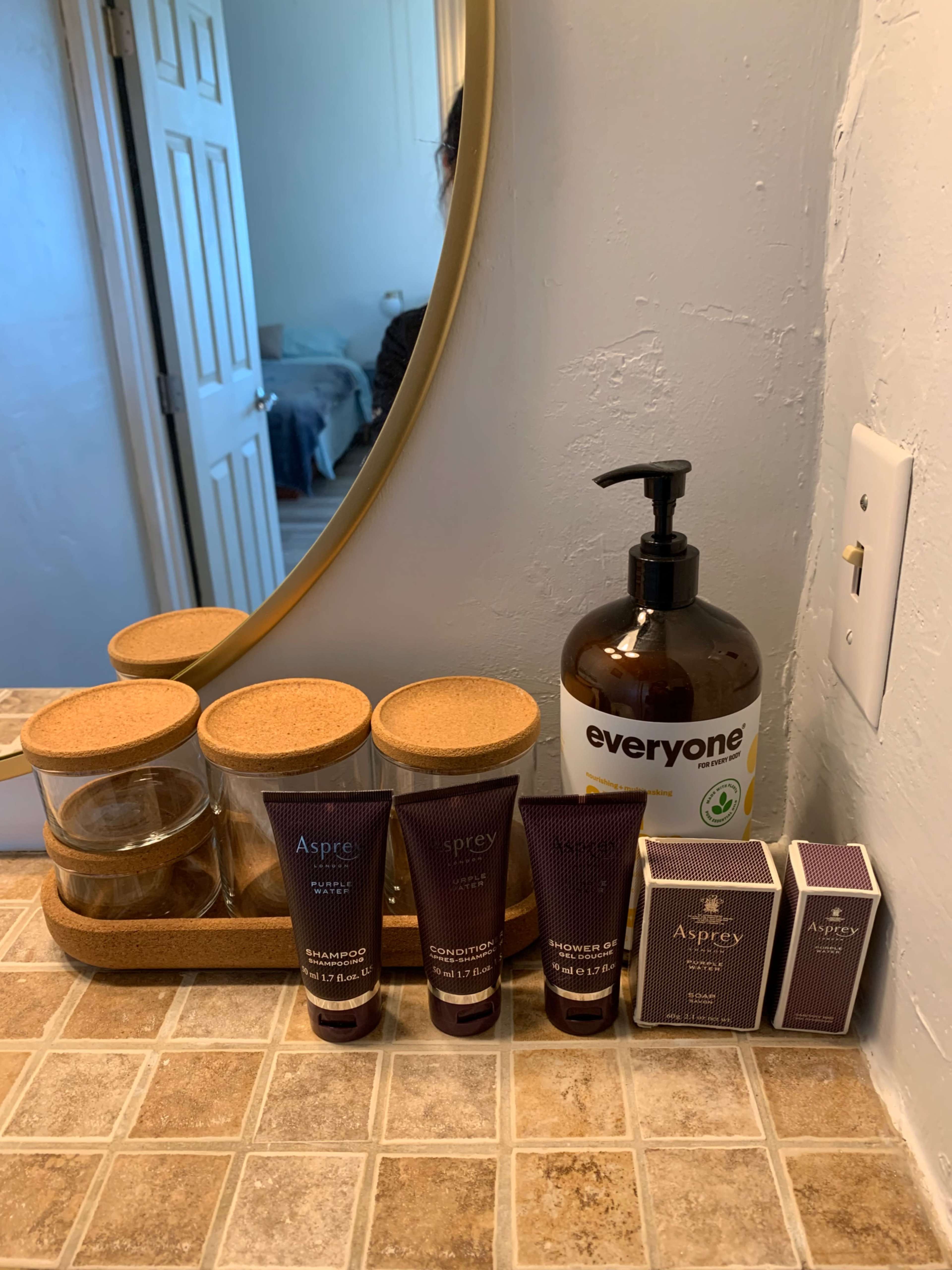 A bathroom countertop featuring skincare products arranged beside a liquid soap dispenser and a mirror reflecting part of a room.