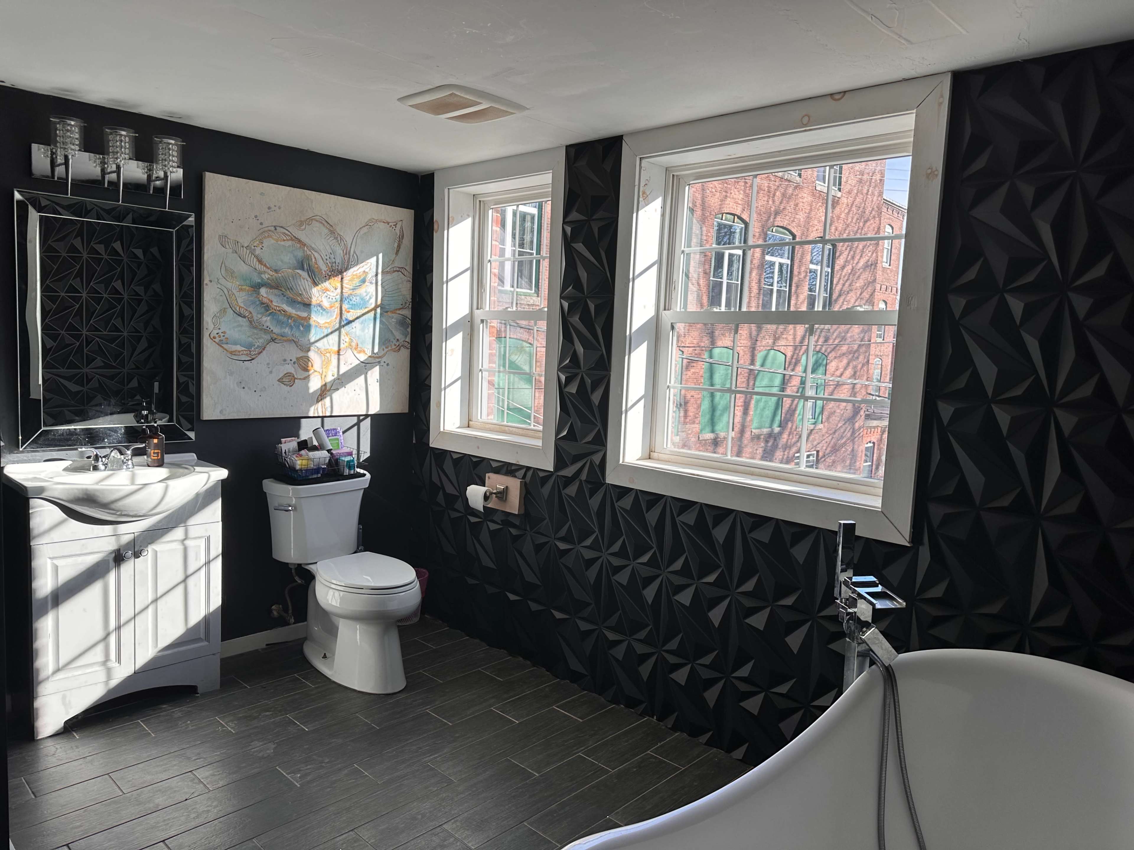 The bathroom features a freestanding bathtub, a toilet, a sink with a mirror, and two large windows illuminating the space.