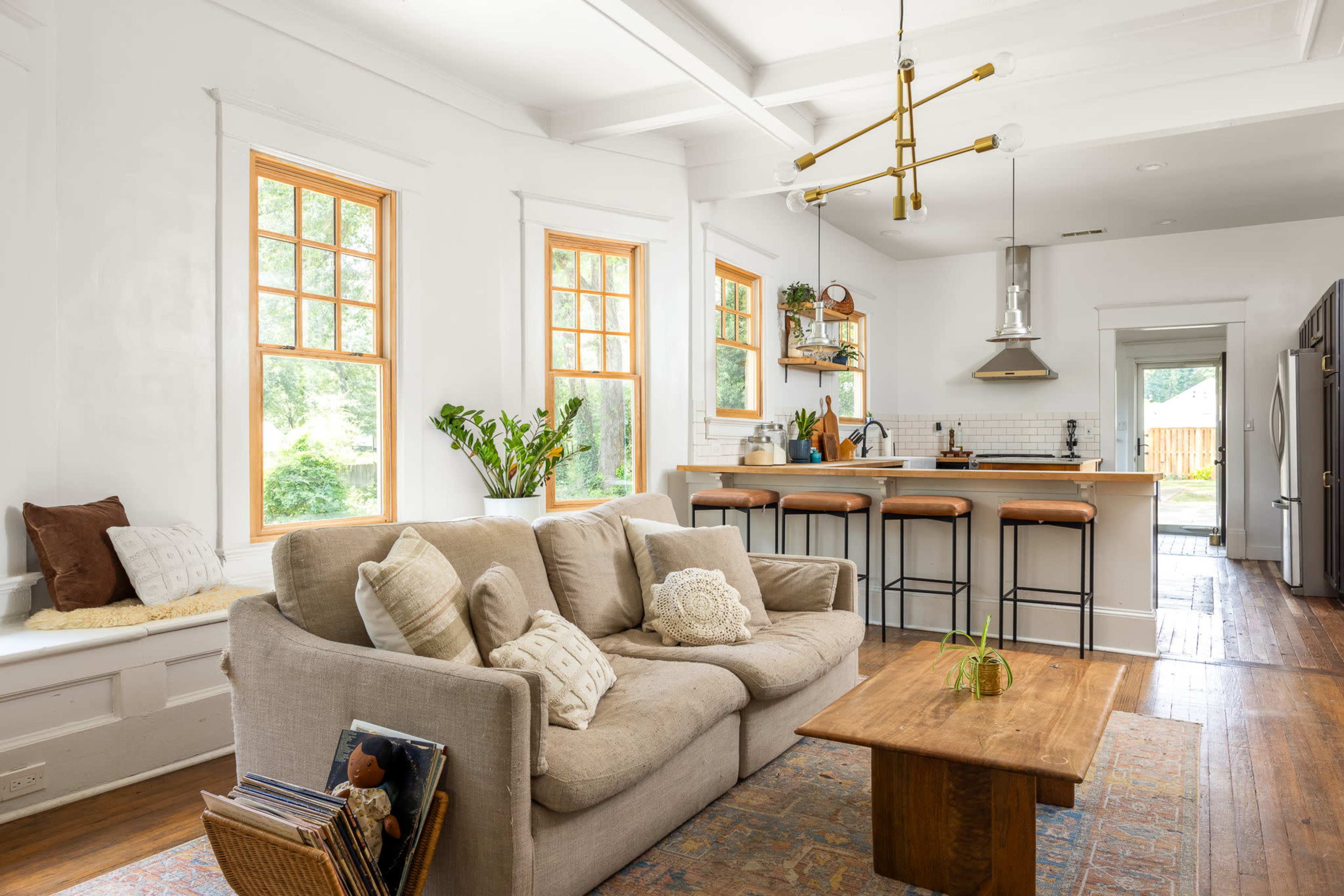 A cozy living room features a beige sofa and a wooden coffee table, with large windows allowing natural light to illuminate the space and a kitchen visible in the background.