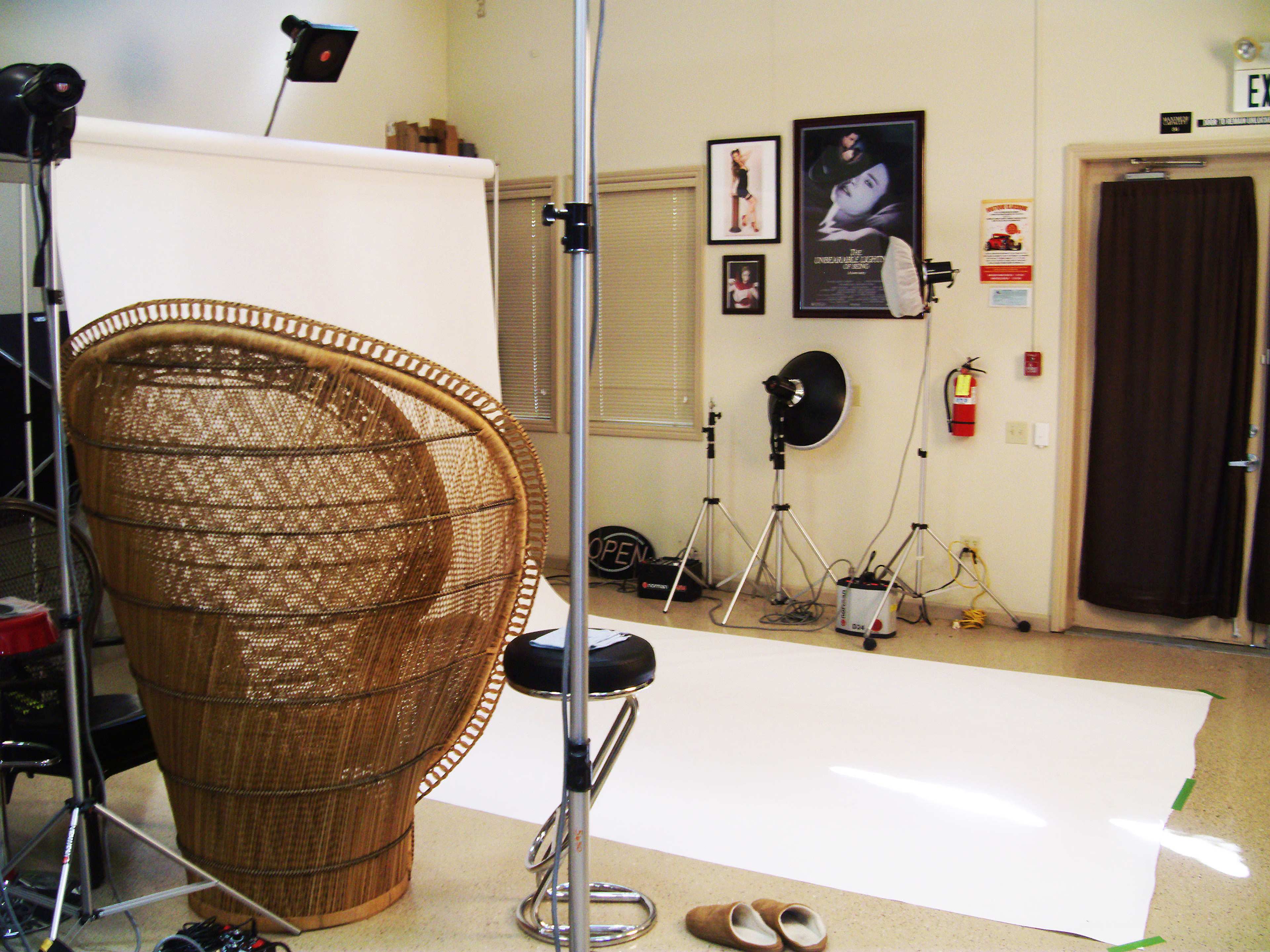 A photography studio with a large wicker chair, a white backdrop, lighting equipment, and a door leading to another area.