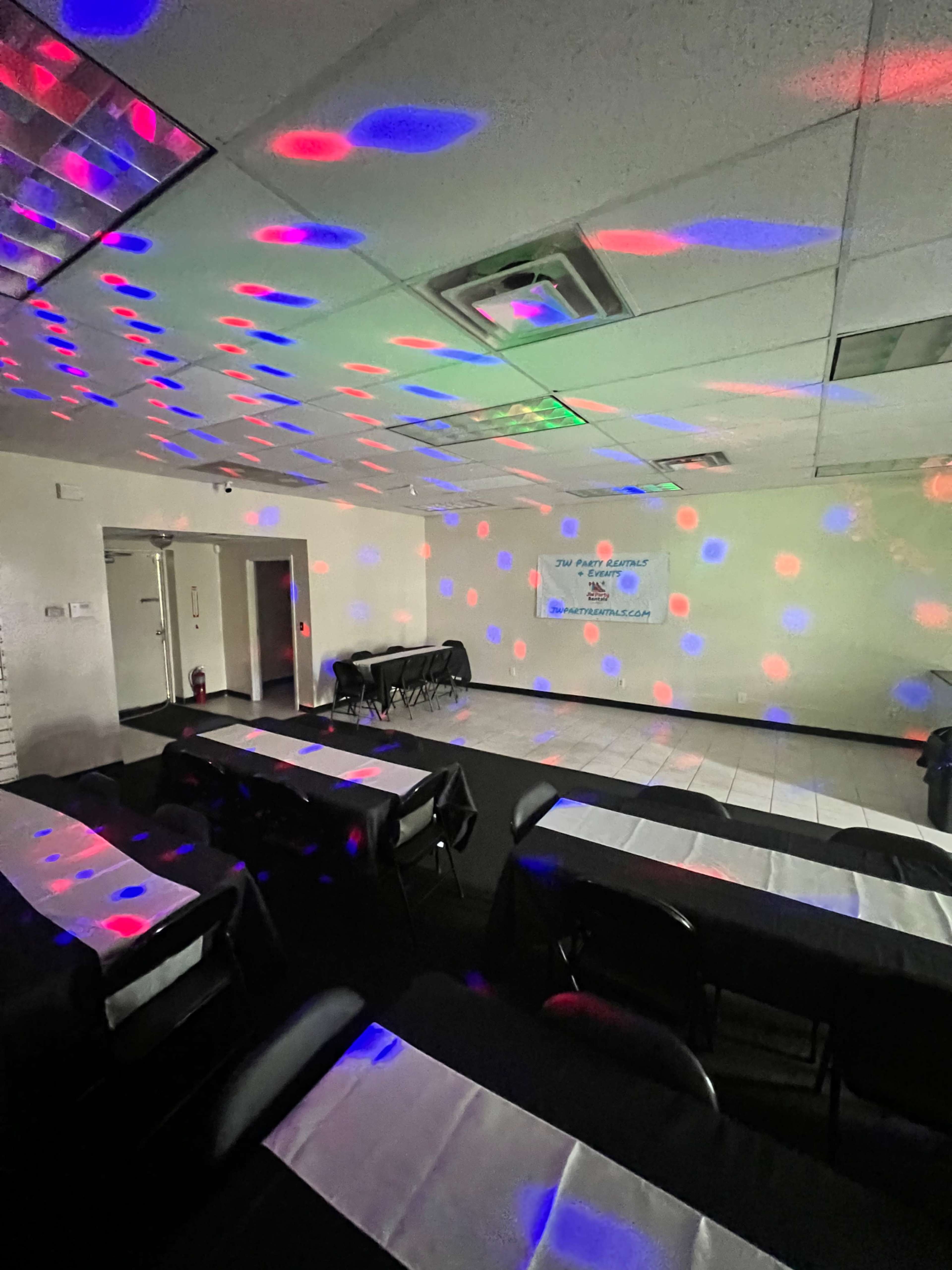 A spacious indoor room features several tables with black tablecloths, illuminated by colored lights creating a festive atmosphere on the walls.