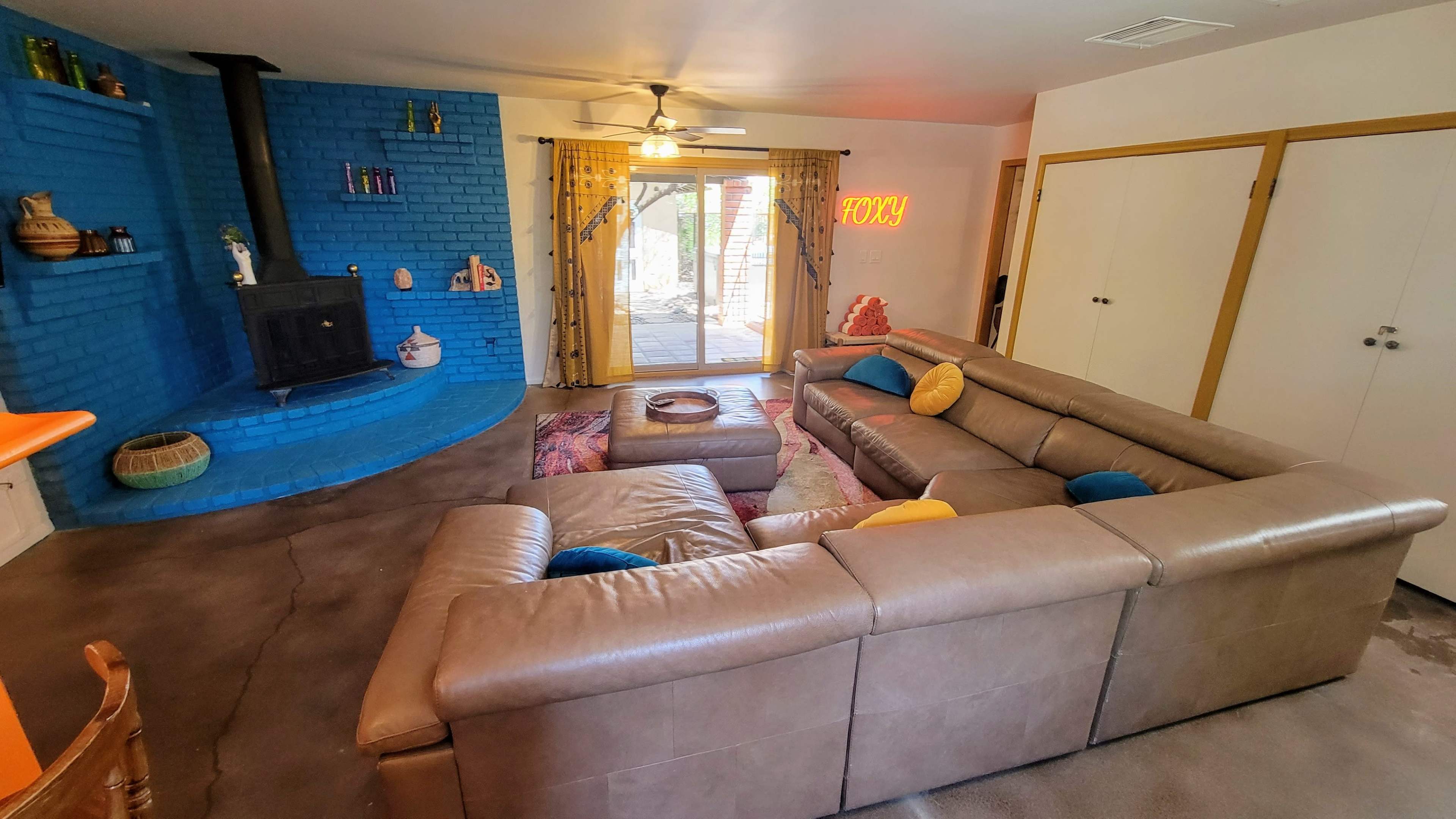 The image shows a living room featuring a light brown sectional sofa, a blue brick wall with decorative items, a wood stove, and a sliding glass door leading to an outdoor area.