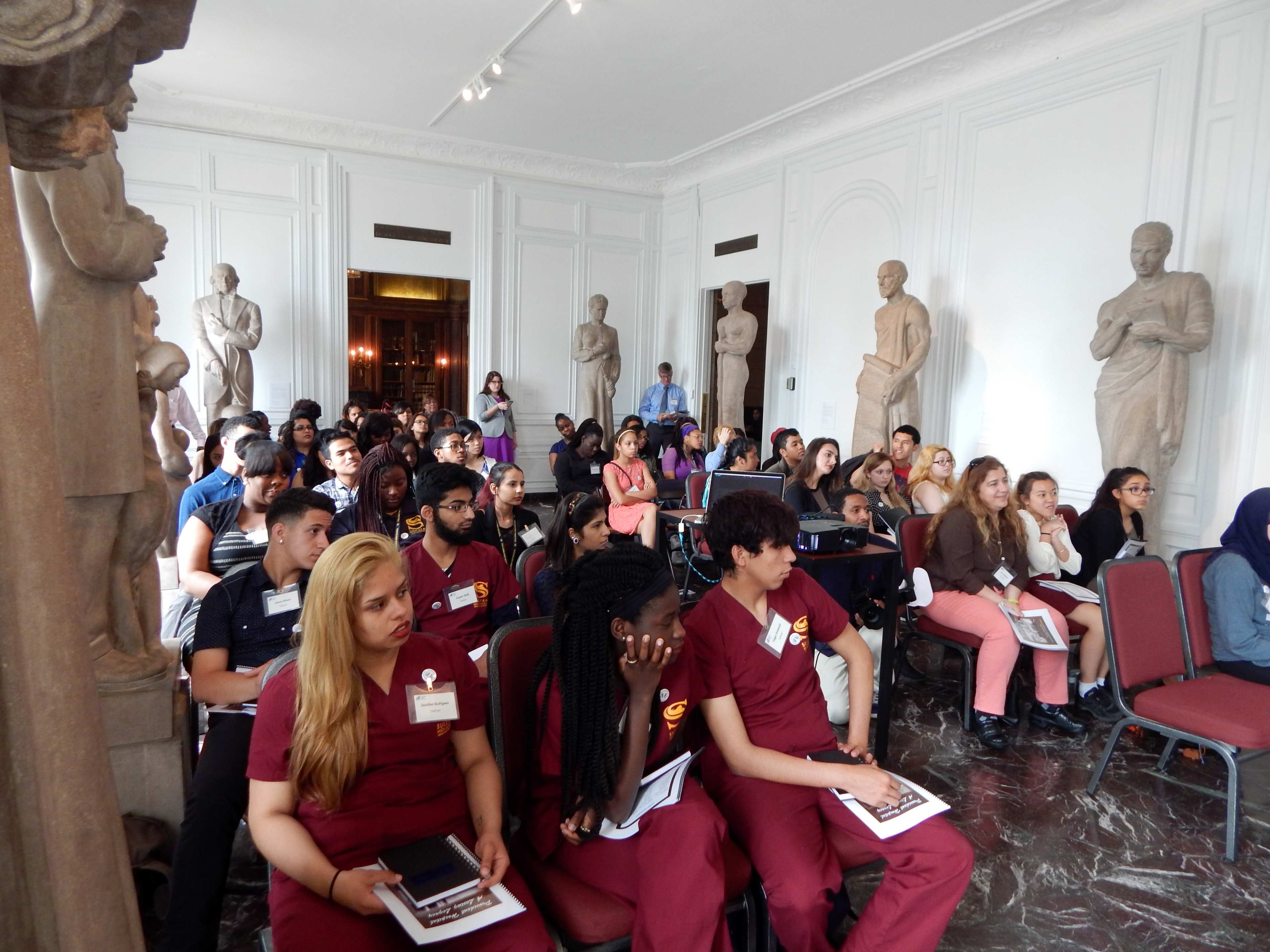 A diverse group of students sits in a spacious room adorned with sculptures, attentively engaged in a presentation.