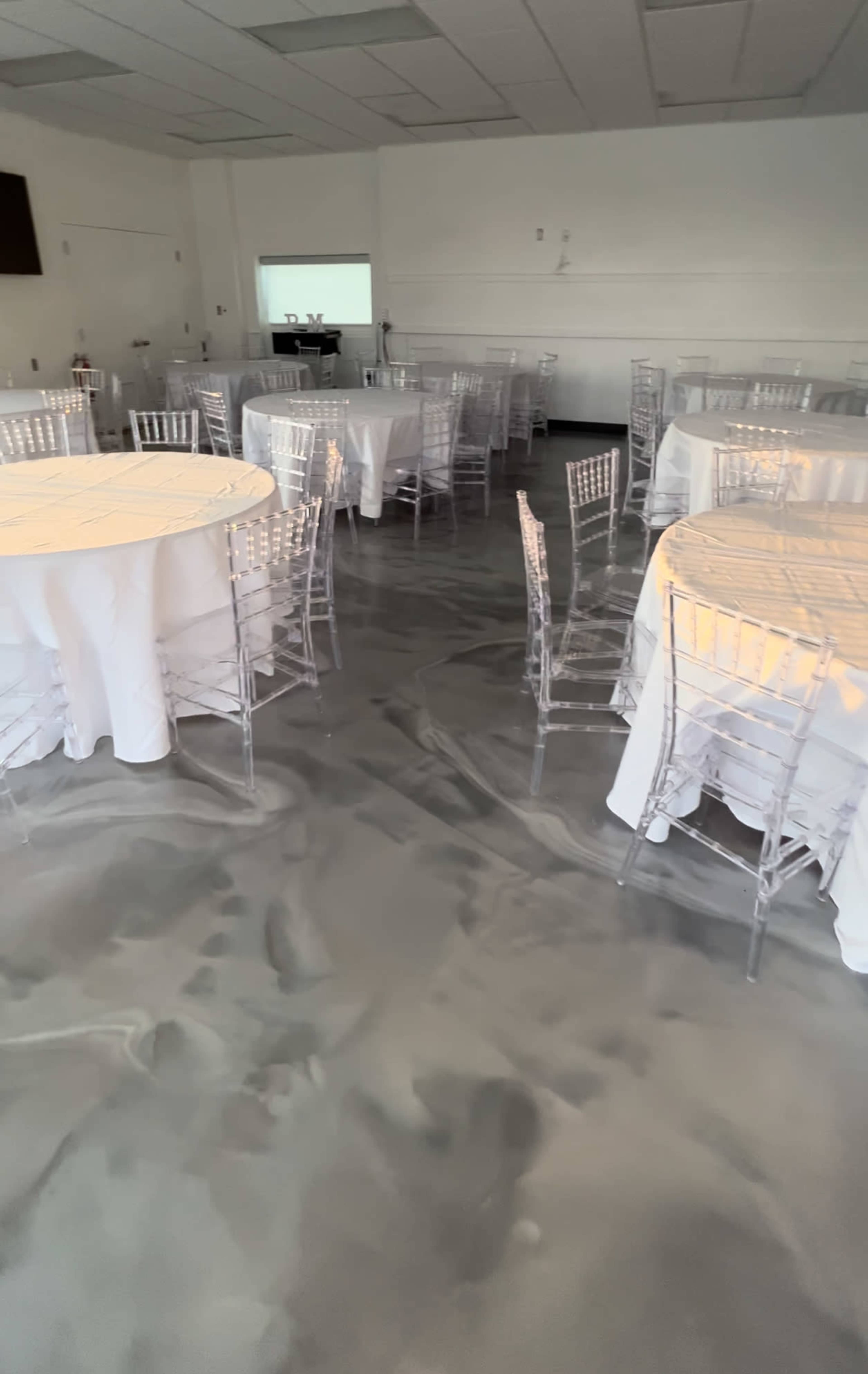 A spacious event area with several round tables covered in white tablecloths and clear acrylic chairs arranged on a marbled gray floor.