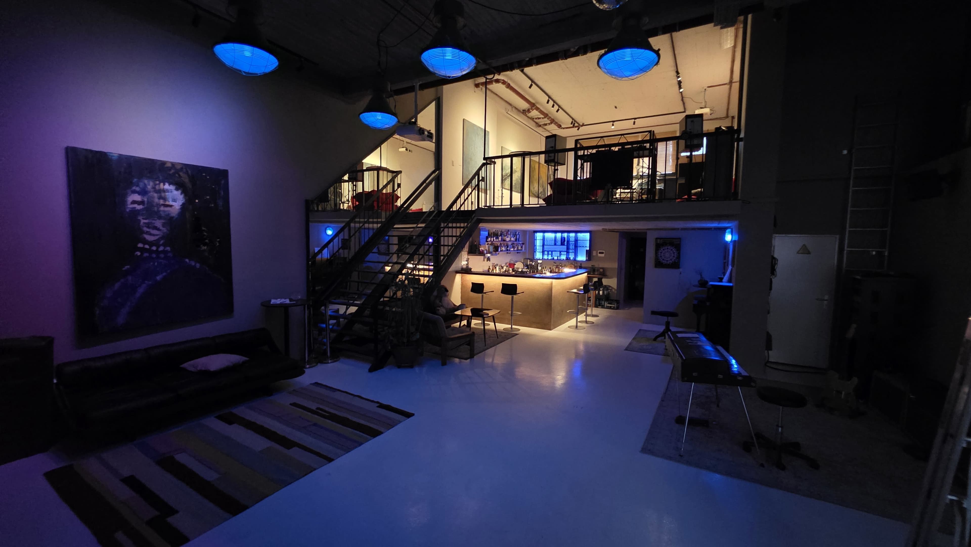 The image shows a spacious, modern loft with a staircase leading to an upper level, featuring a bar area illuminated by blue lights.