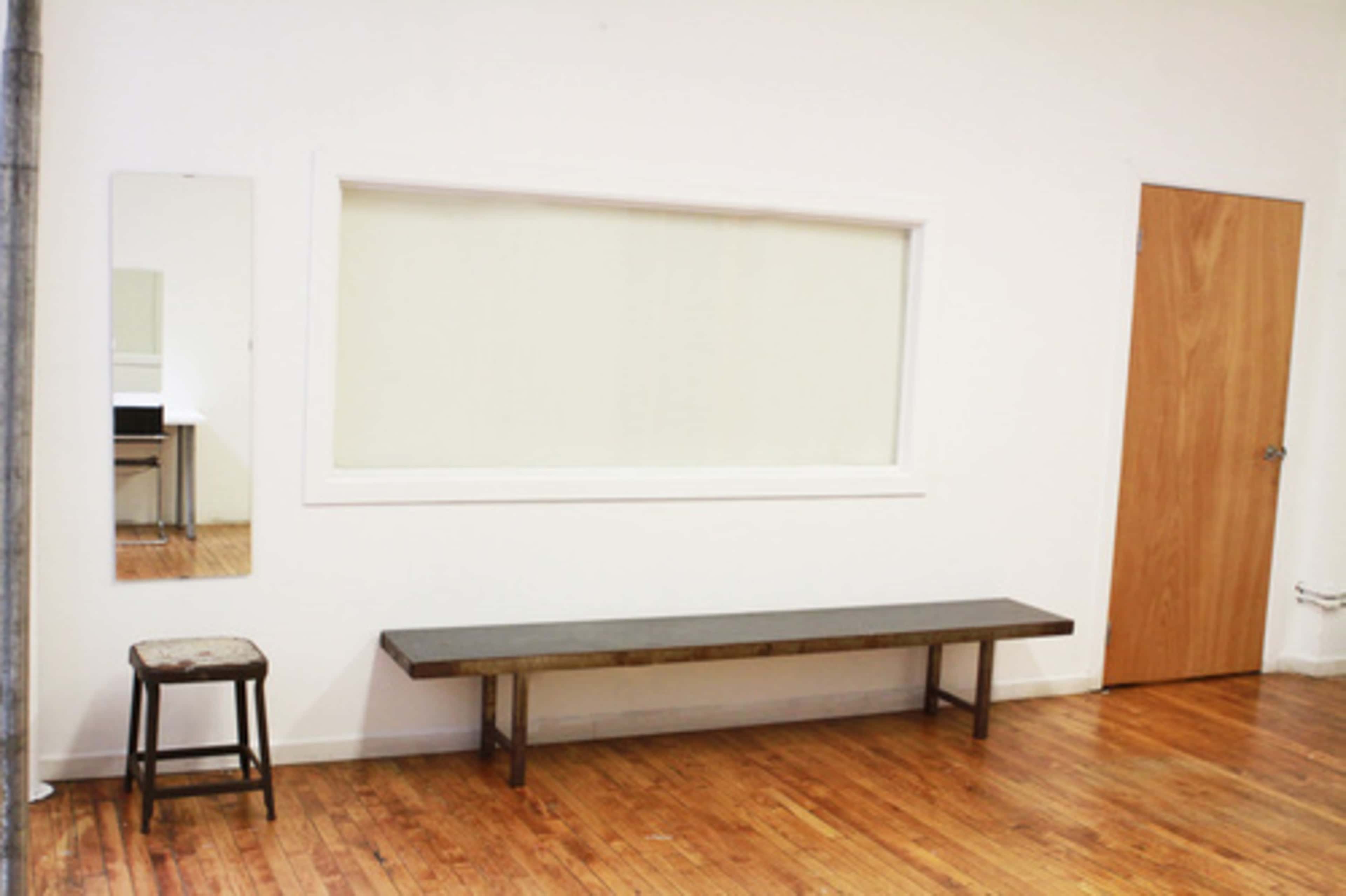 A minimalist room with a wooden bench, a small stool, a large window, and a mirror on the wall.