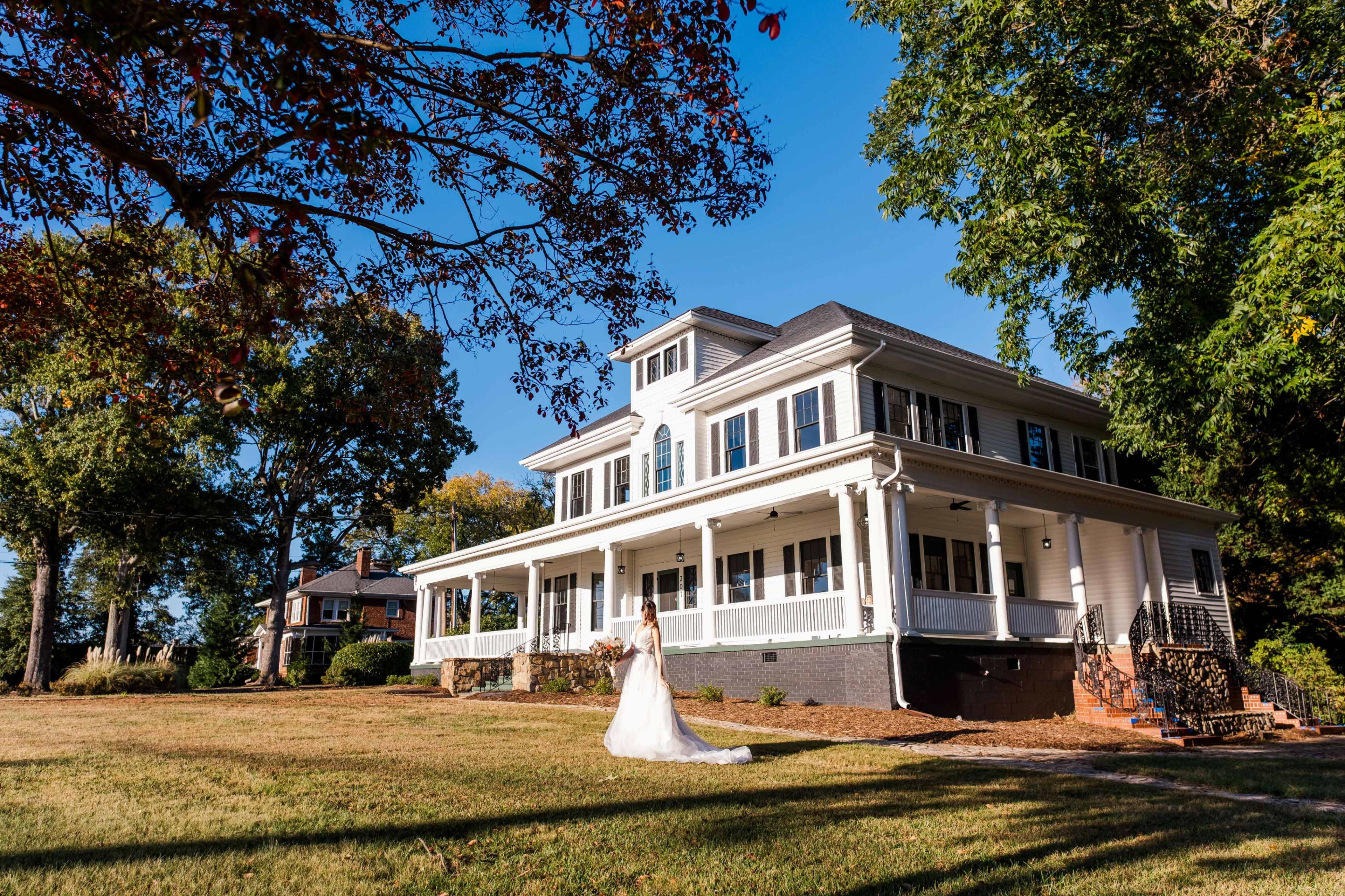 A bride in a white wedding dress walks across the lawn of a large, two-story house with a porch and blue shutters.