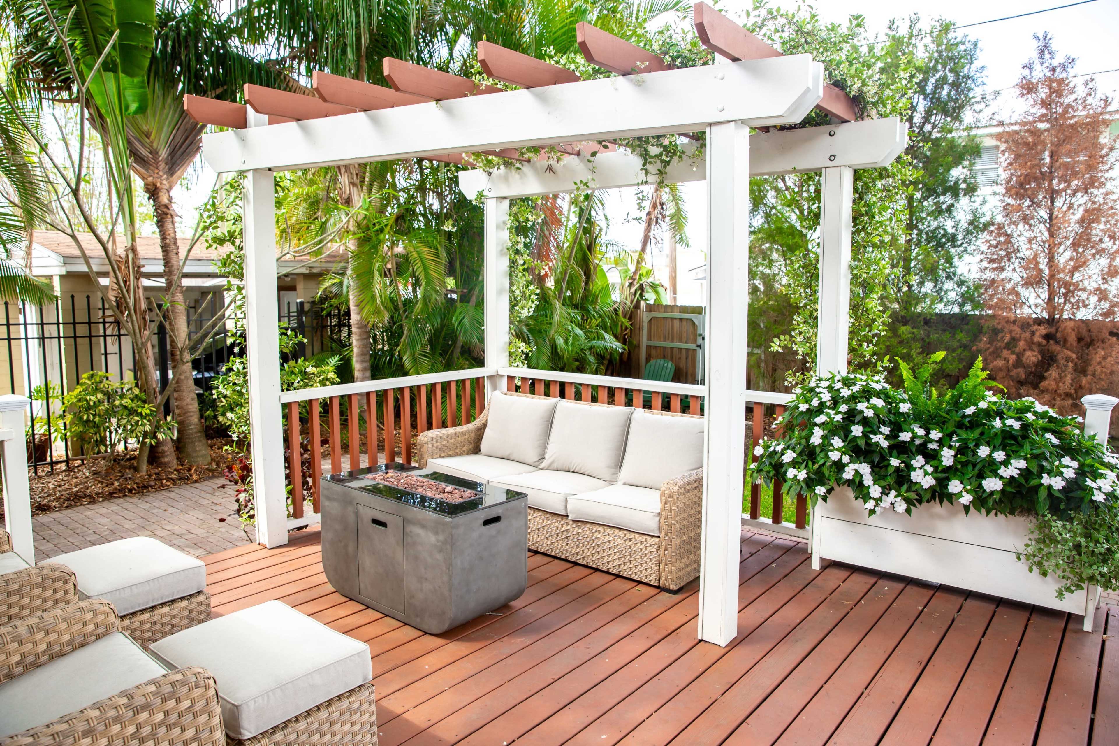 The image shows a patio with a pergola, featuring a seating area with cushions and a fire pit, surrounded by greenery.