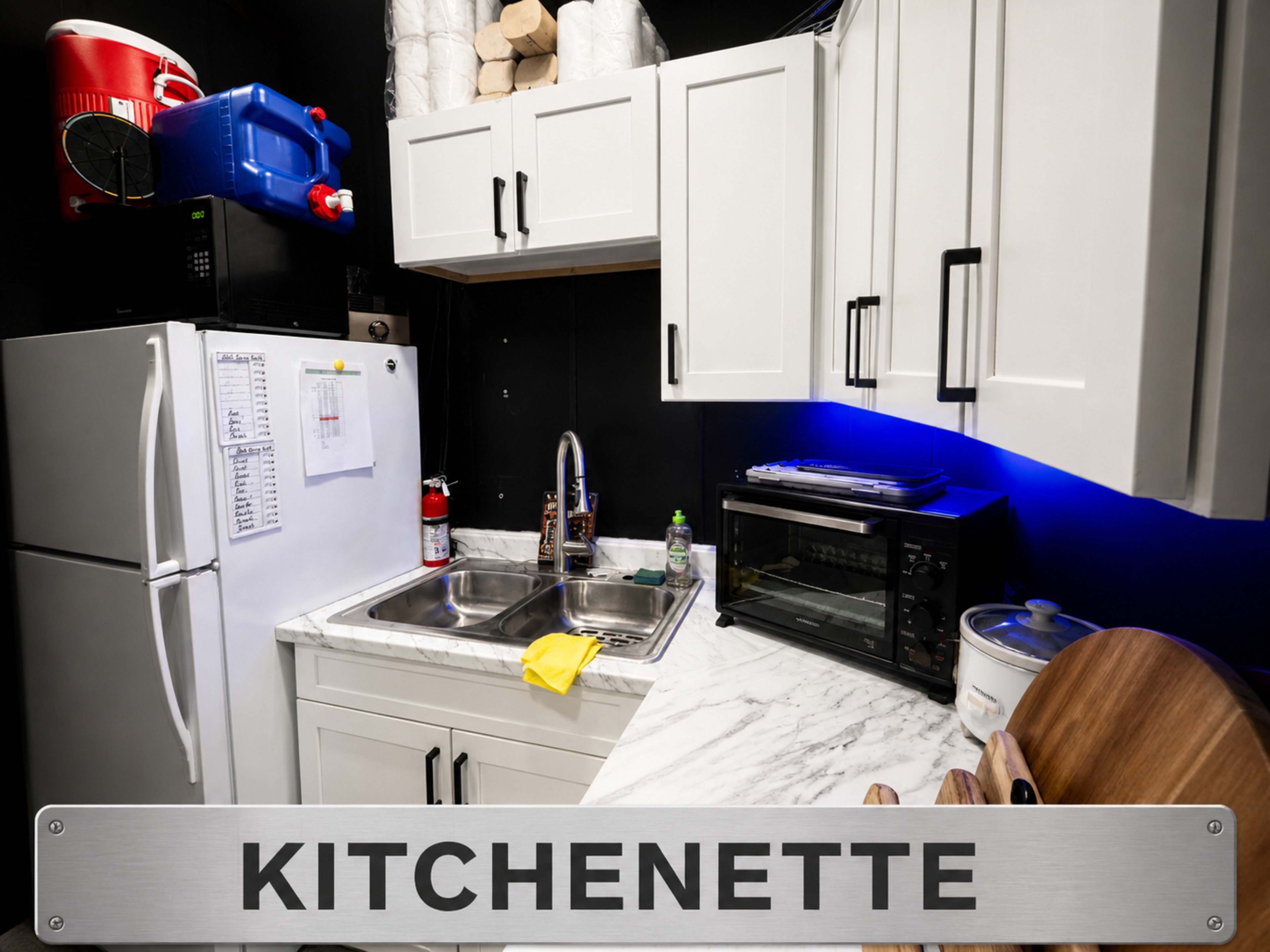 The image shows a small kitchenette featuring a refrigerator, a sink, and a microwave, with cabinetry above and items stored nearby.