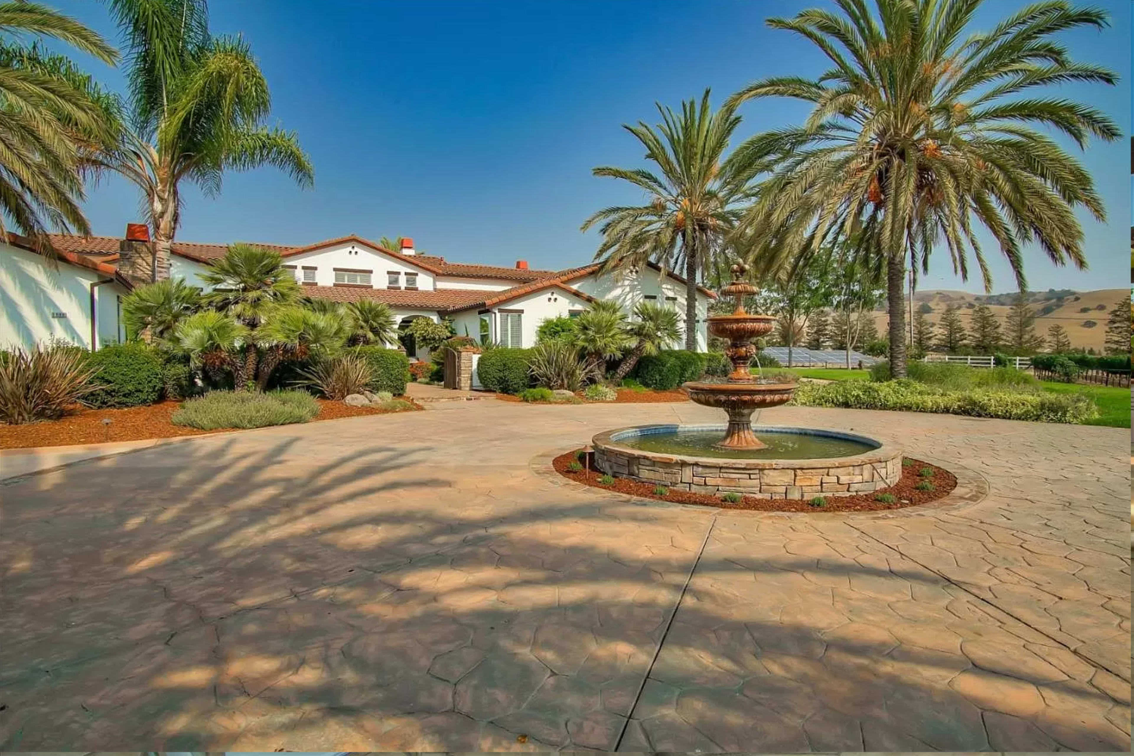 A landscaped courtyard with a fountain surrounded by palm trees and a large house in the background.