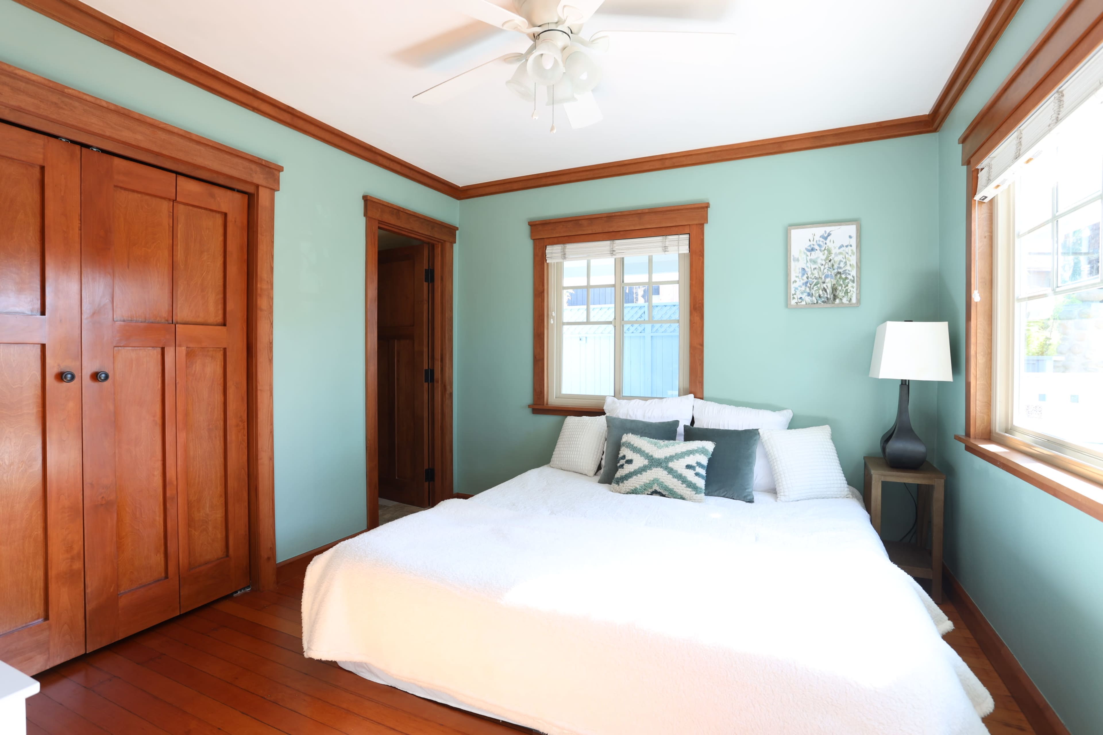 The image shows a simple bedroom featuring a bed with white bedding, a decorative pillow, a nightstand with a lamp, and wooden closet doors.