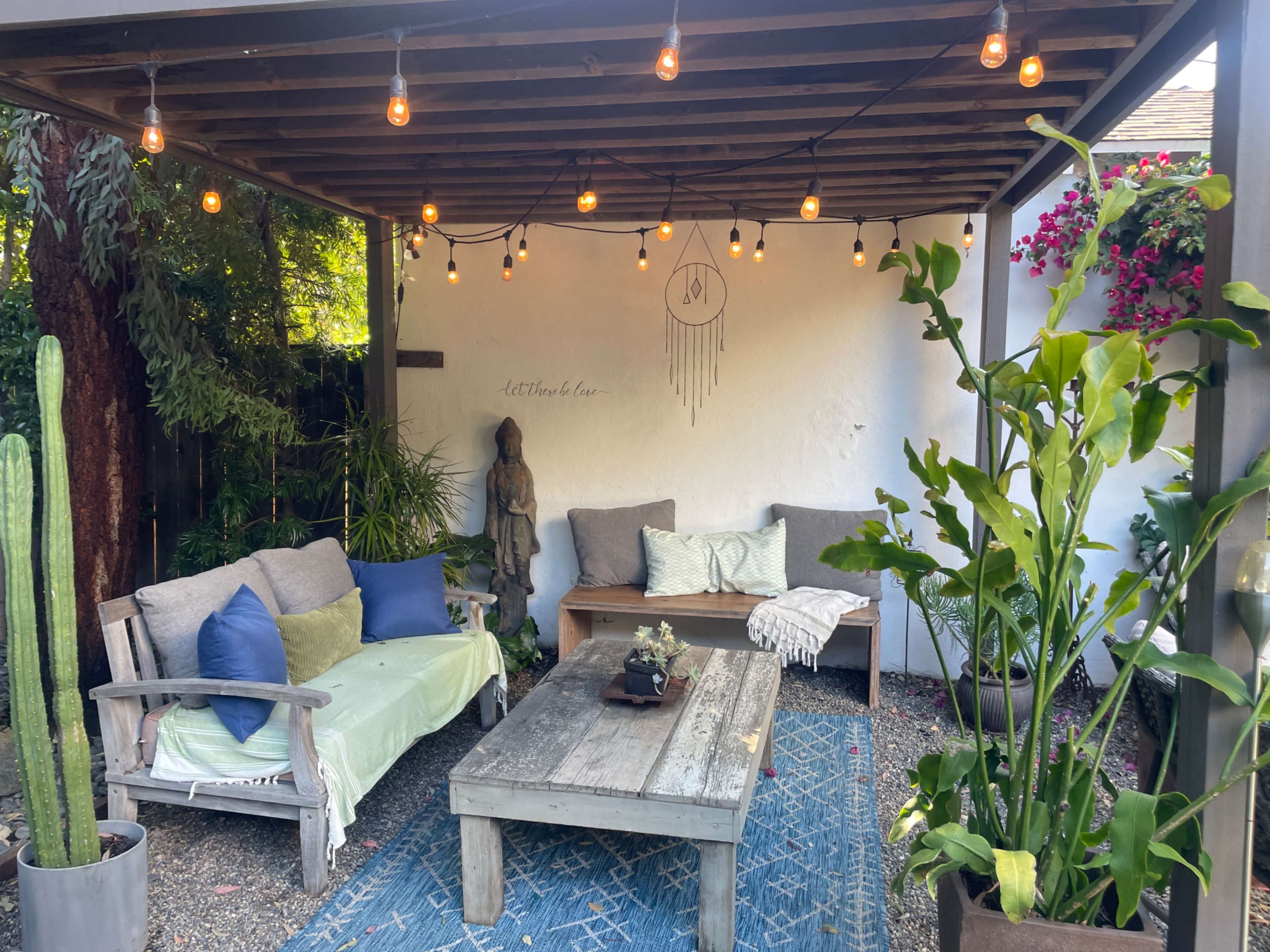 A cozy outdoor patio features string lights overhead, a wooden coffee table, seating with cushions, potted plants, and a decorative statue against a white wall.