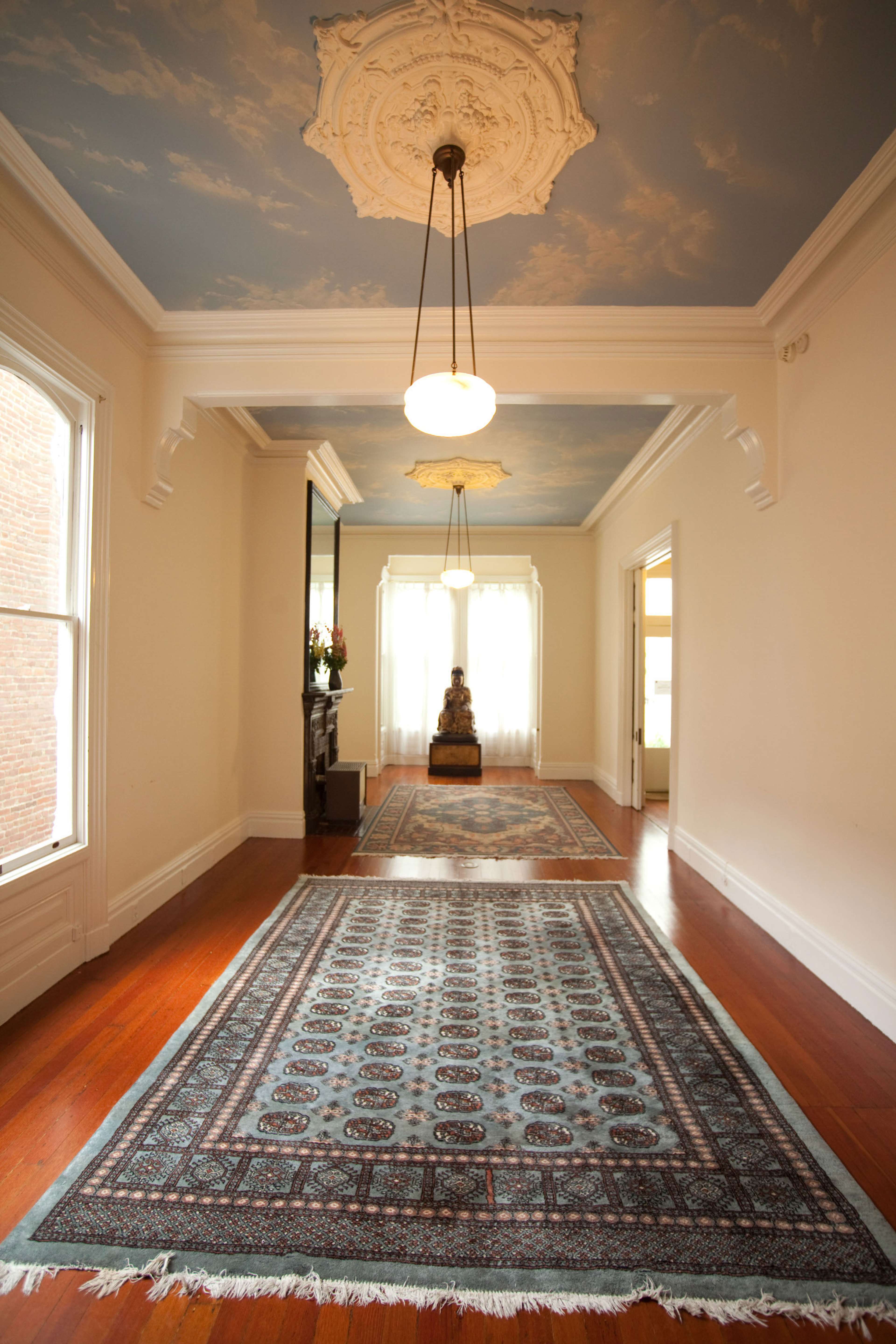 The hallway features a patterned rug, ornate ceiling details, and soft lighting, leading to a room with a statue and large windows.