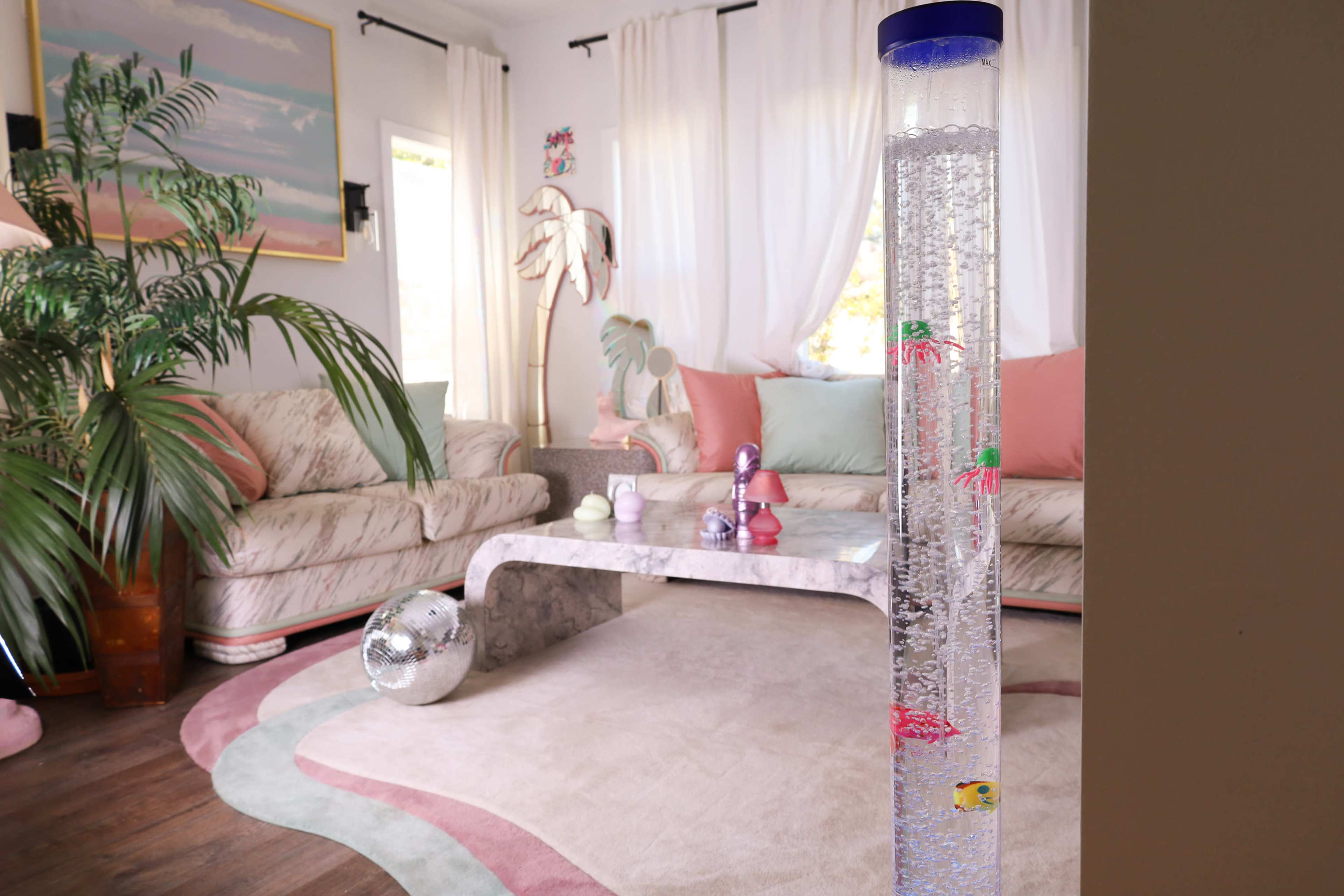 A tall, cylindrical aquarium filled with water and colorful fish stands in a living room featuring a patterned rug and a light-colored sofa.