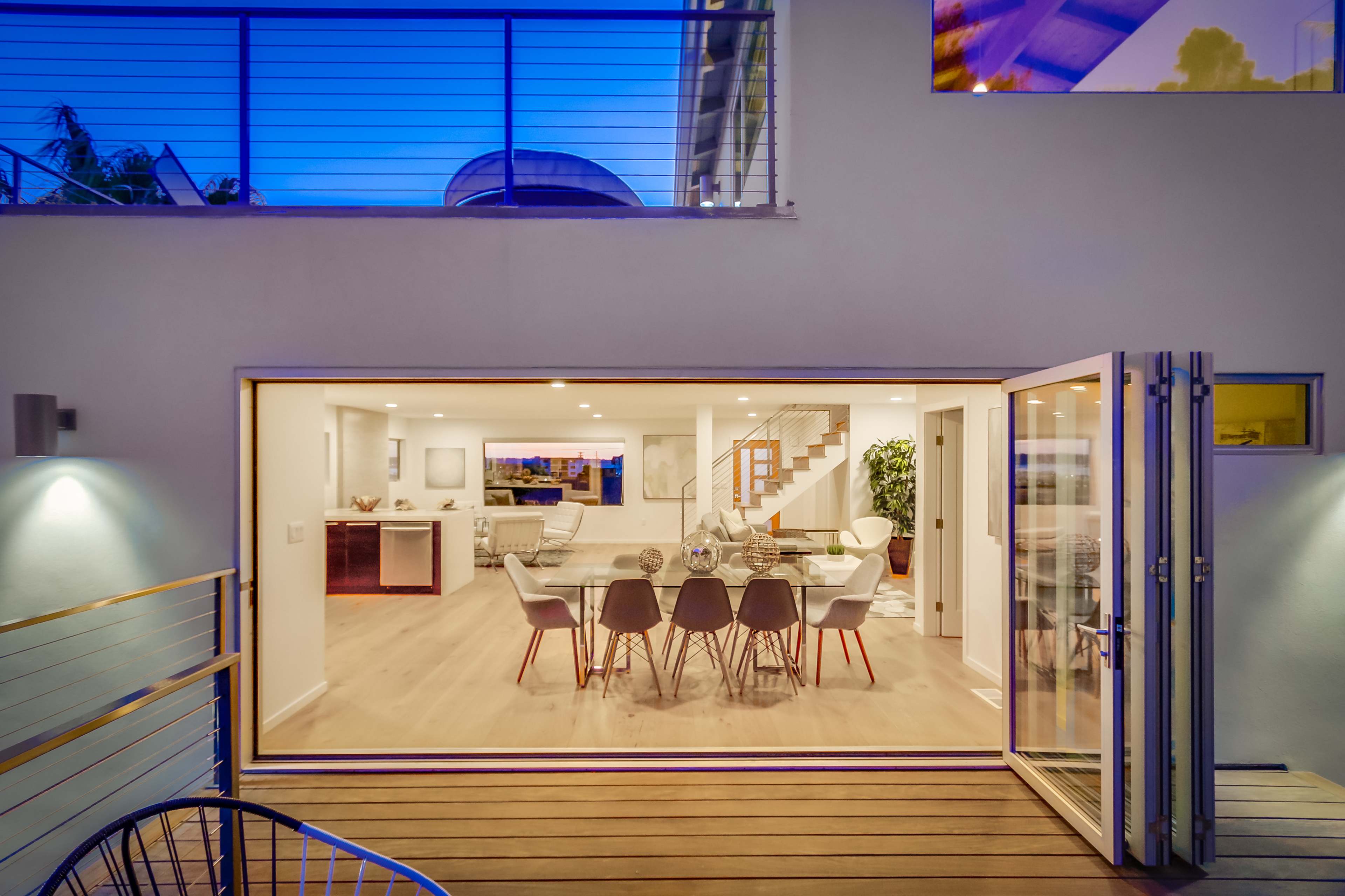 The image shows a modern dining area with a large table and chairs, seen through open sliding glass doors, leading to a wooden deck and a staircase in the background.