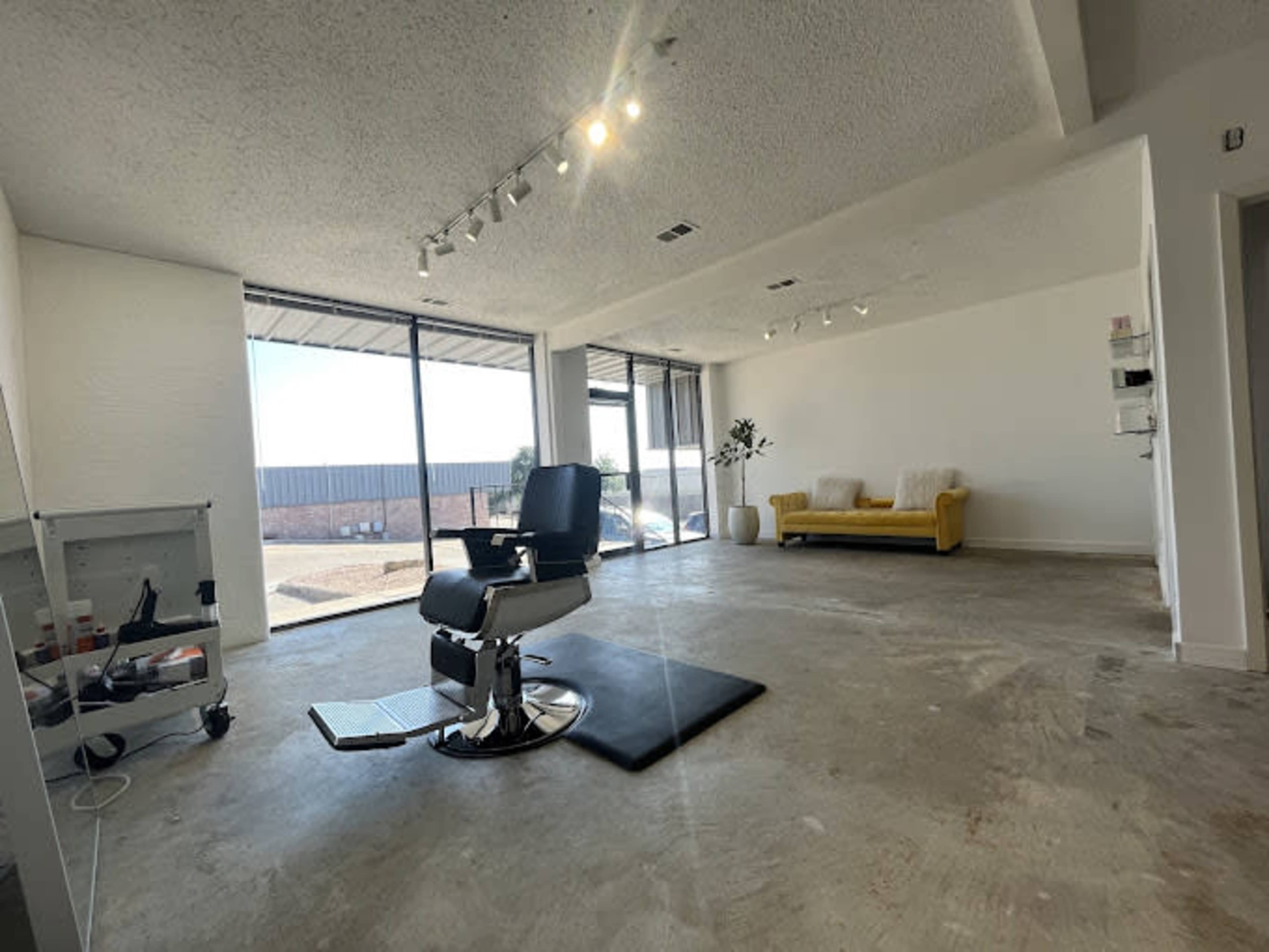 The image shows an empty, modern barber shop with a black barber chair, large windows letting in natural light, and a yellow couch against a white wall.