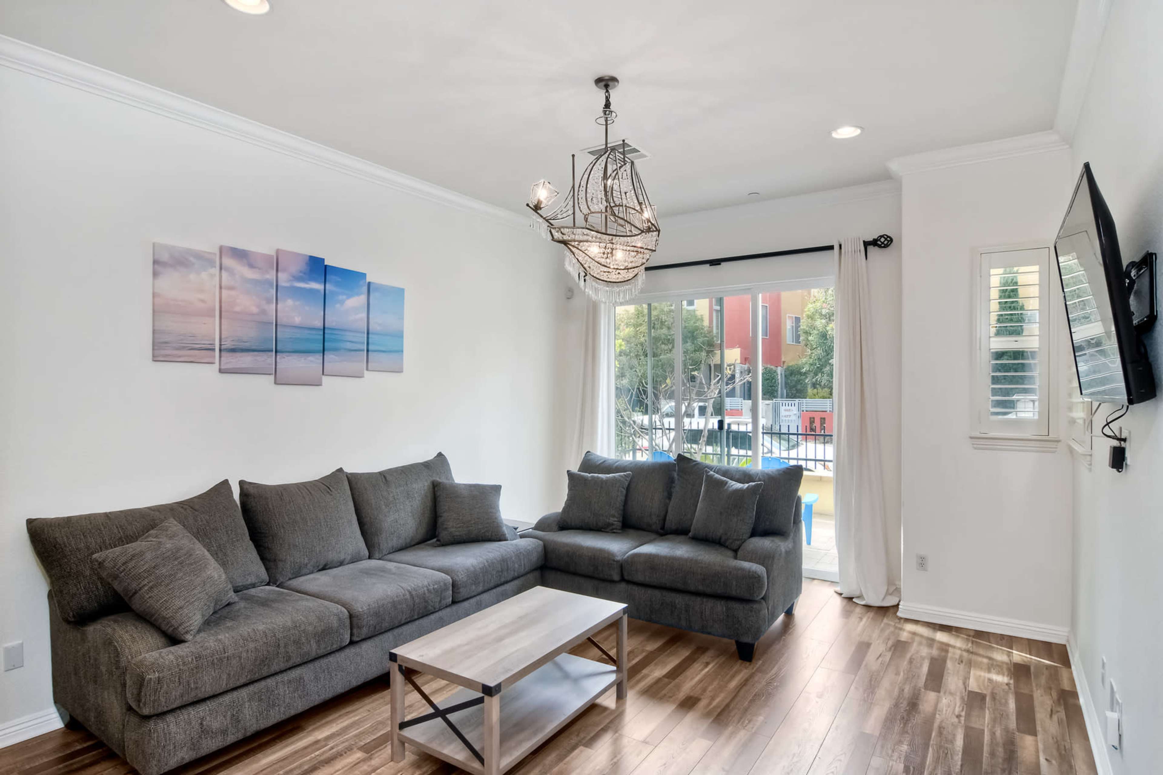 A modern living room features a gray sectional sofa, a light wooden coffee table, and a decorative chandelier, with large windows overlooking an outdoor space.