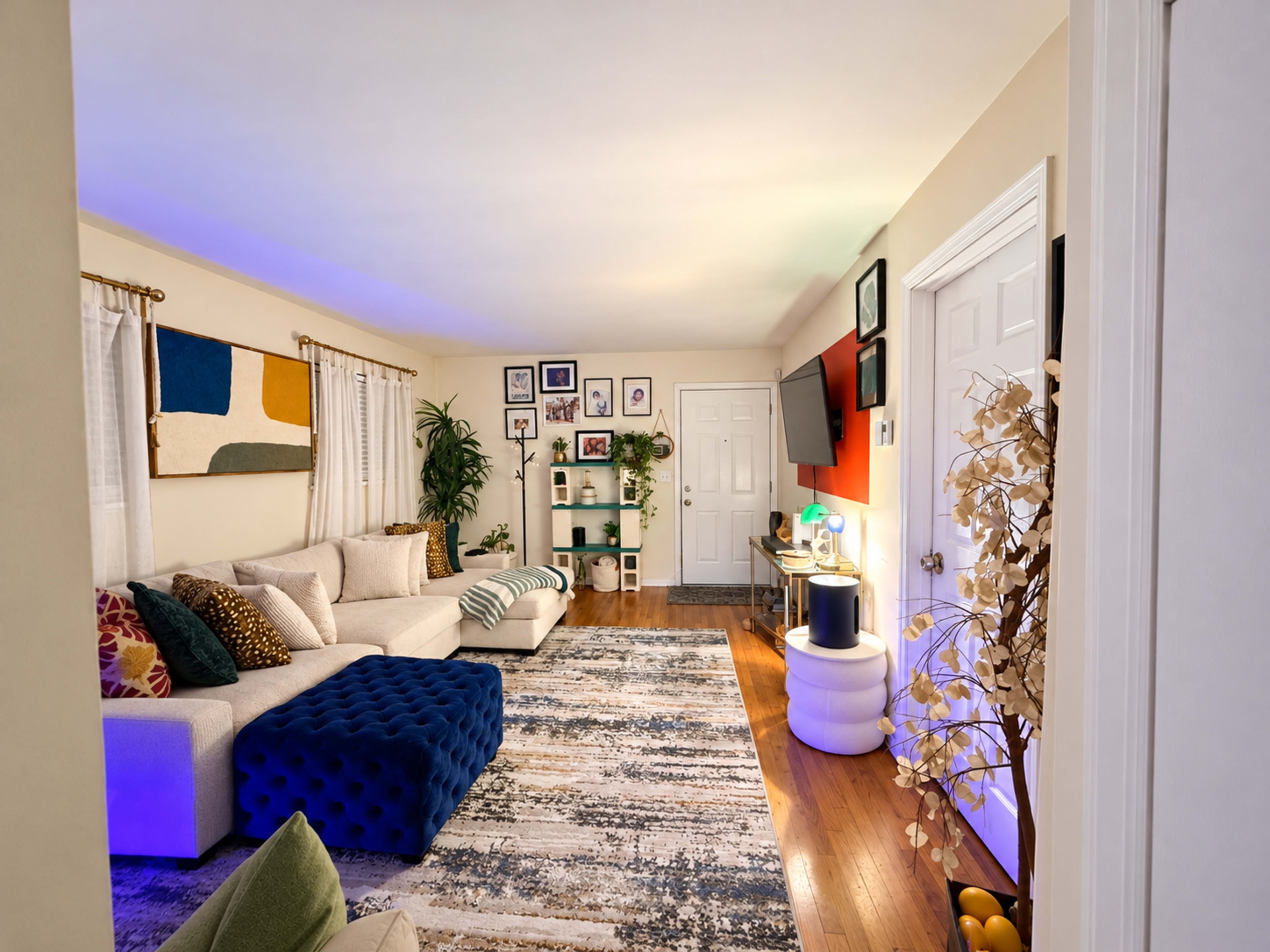 The image shows a well-decorated living room featuring a large sectional sofa, a blue tufted ottoman, and a patterned area rug, with various wall art and plants enhancing the space.
