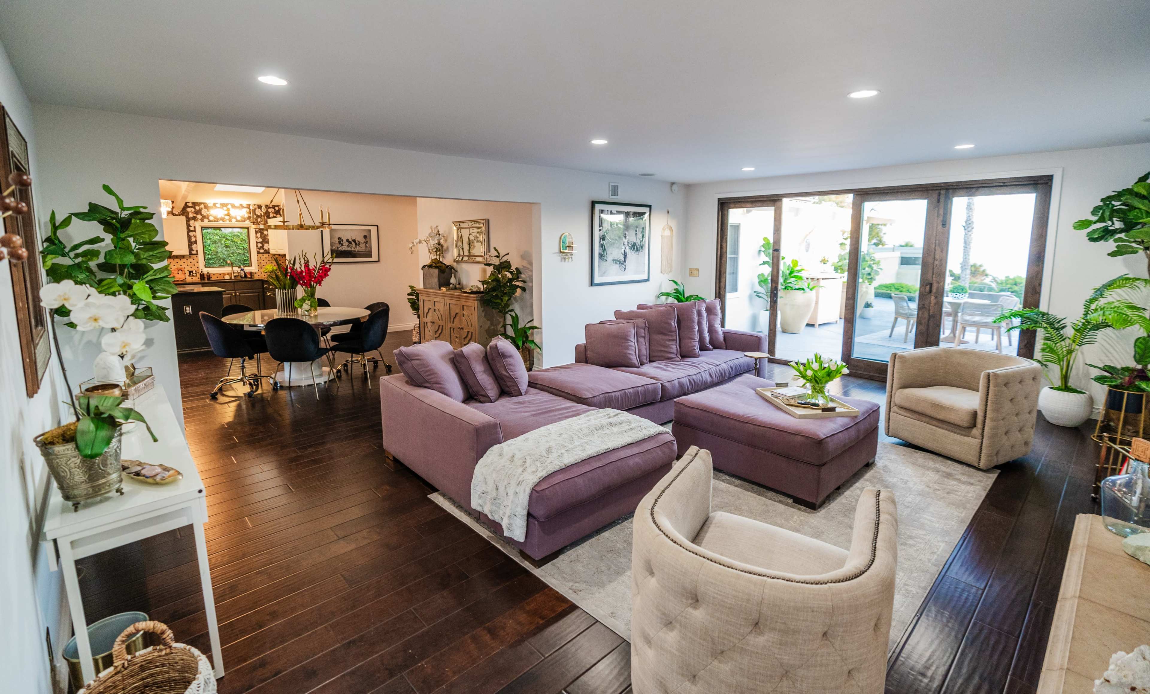 The living room features a spacious layout with a large purple sectional sofa, wooden flooring, and plants, leading to a patio area through sliding glass doors.