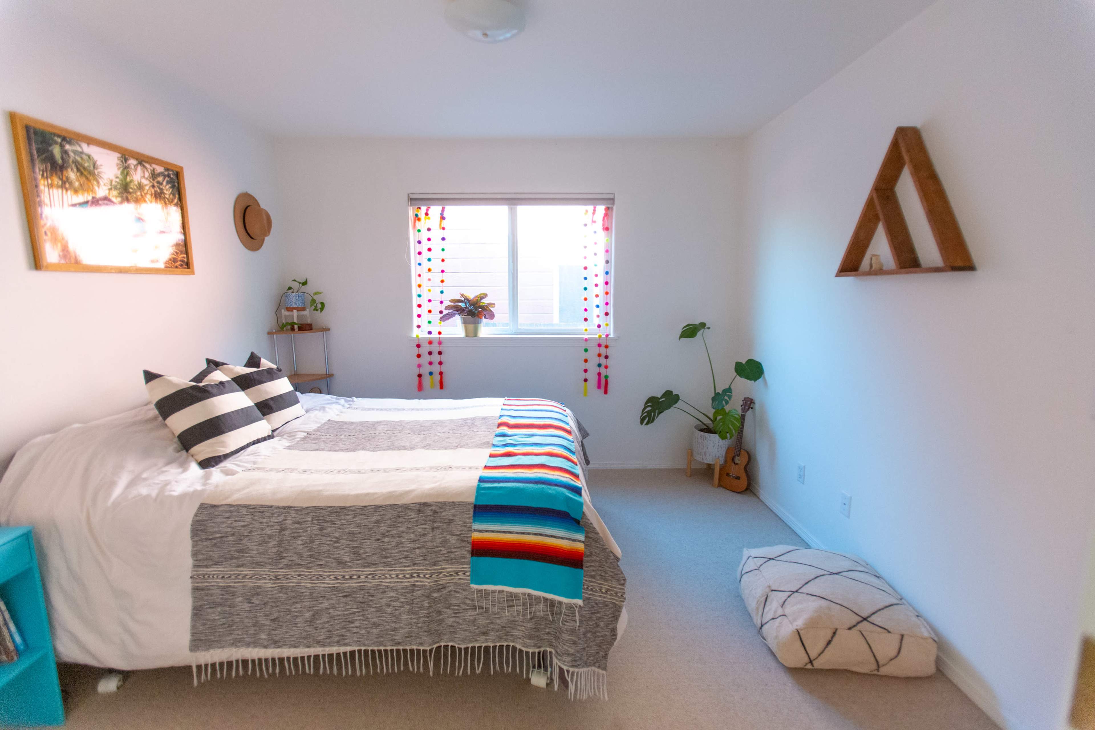 The room features a bed with patterned blankets, a window adorned with colorful curtains, and a small plant next to a guitar on the floor.