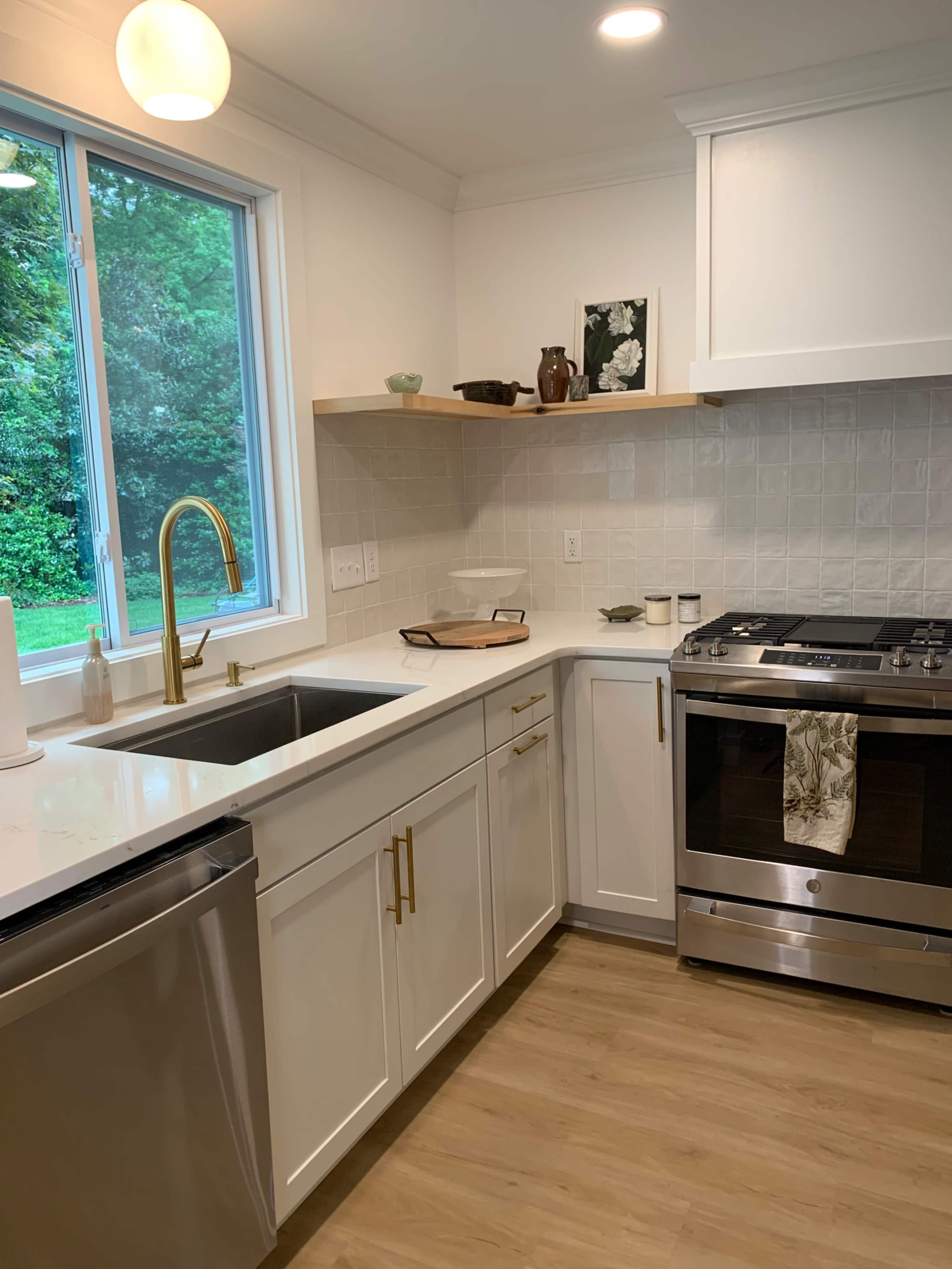 A modern kitchen features white cabinetry, a stainless steel stove, and a large window overlooking a garden.