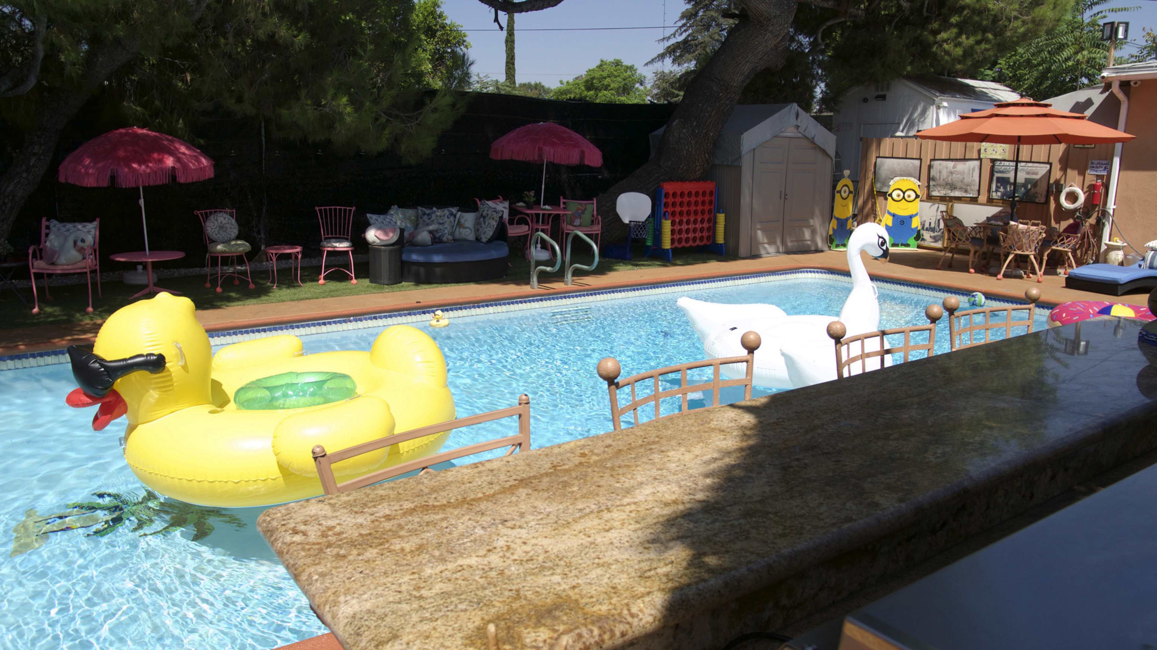 The scene features a backyard pool with inflatable yellow duck and white swan floats, surrounded by patio furniture, umbrellas, and a children's play area.